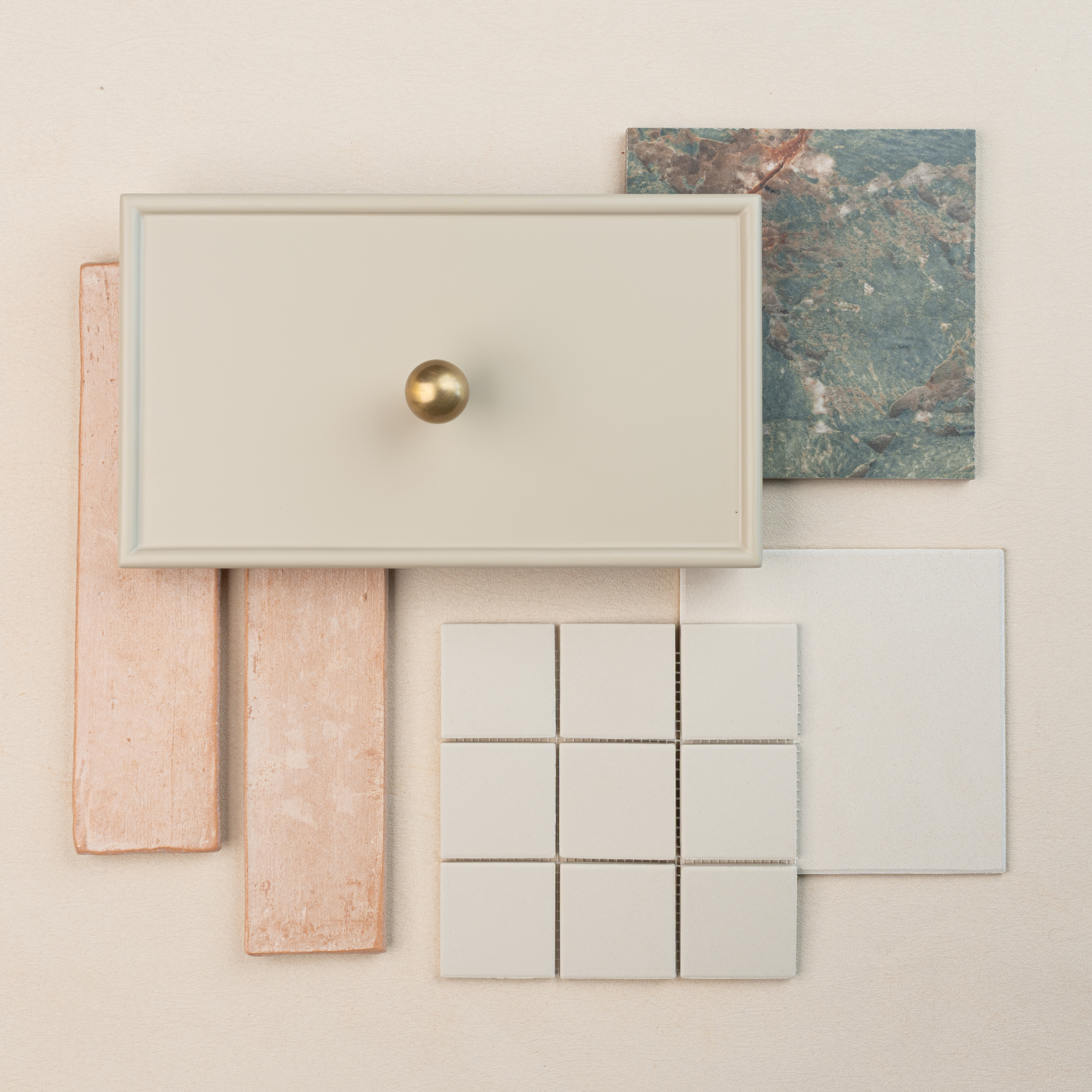 Flat lay of cream cabinet drawer front with brass knob, pink rectangular tiles, green marble tile, and white square ceramic tiles on pale background