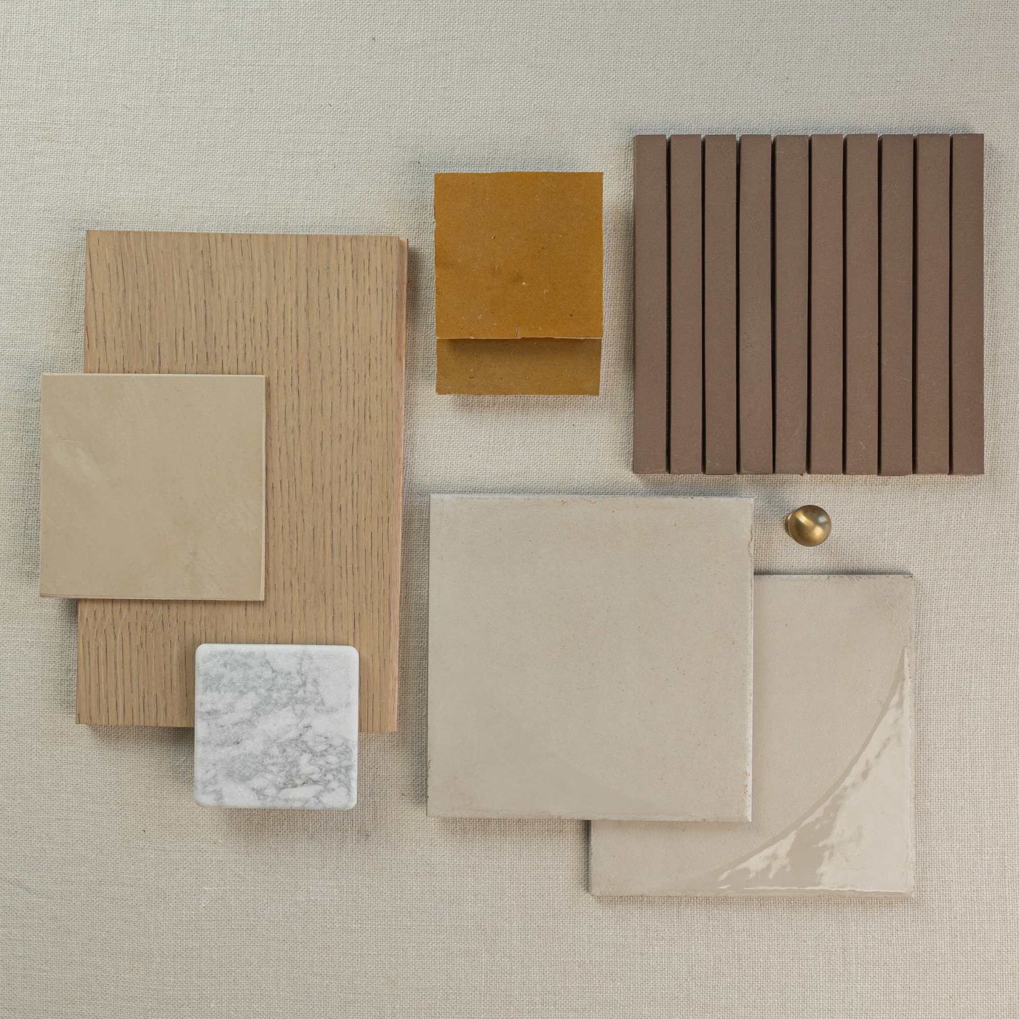 Flat arrangement of beige ceramic and porcelain tiles, mustard and taupe tiles, oak wood sample, marble tile, and brass knob on neutral fabric background
