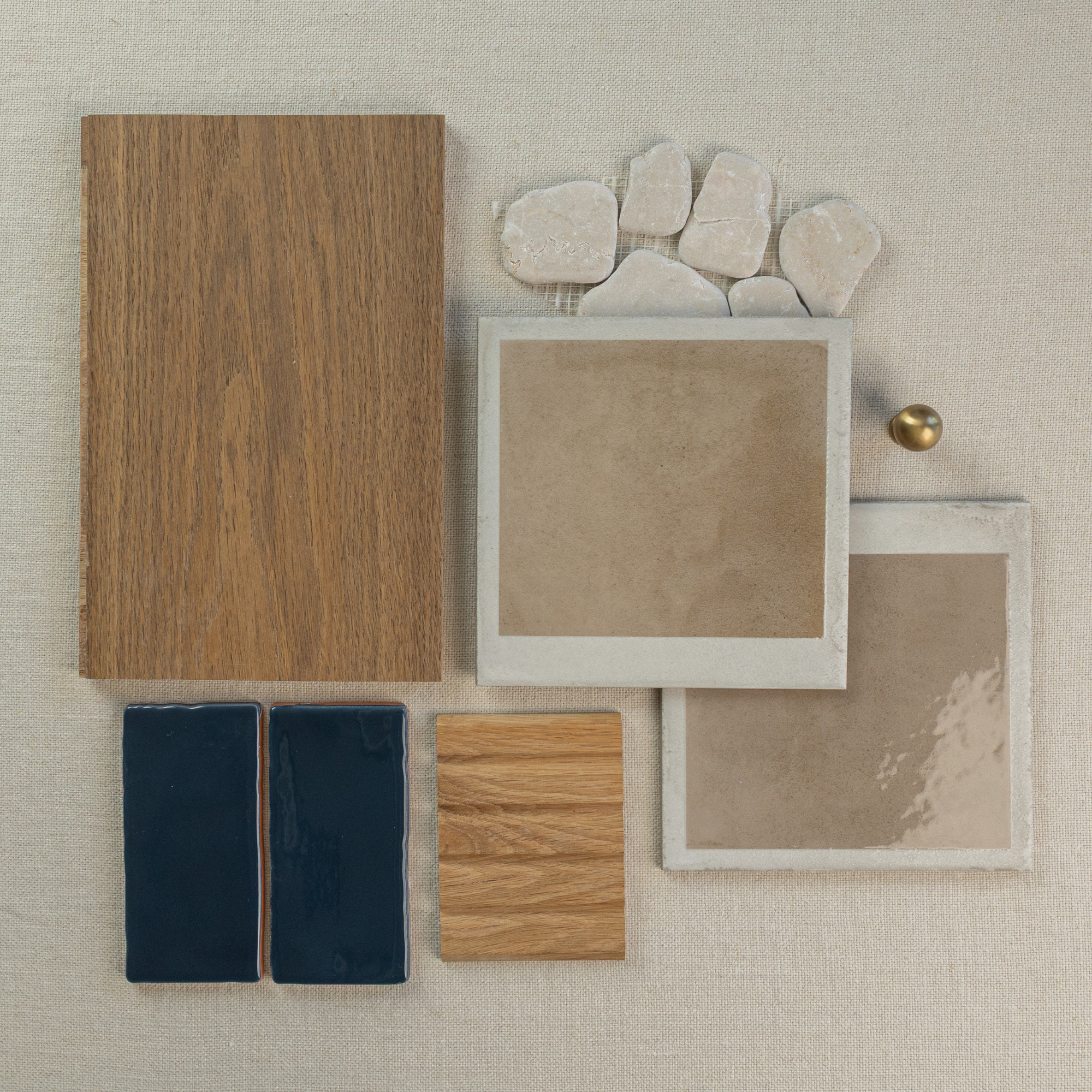 Flat arrangement of warm wood samples, beige porcelain tiles, natural stone pieces, dark blue ceramic tiles, and brass knob on light fabric background