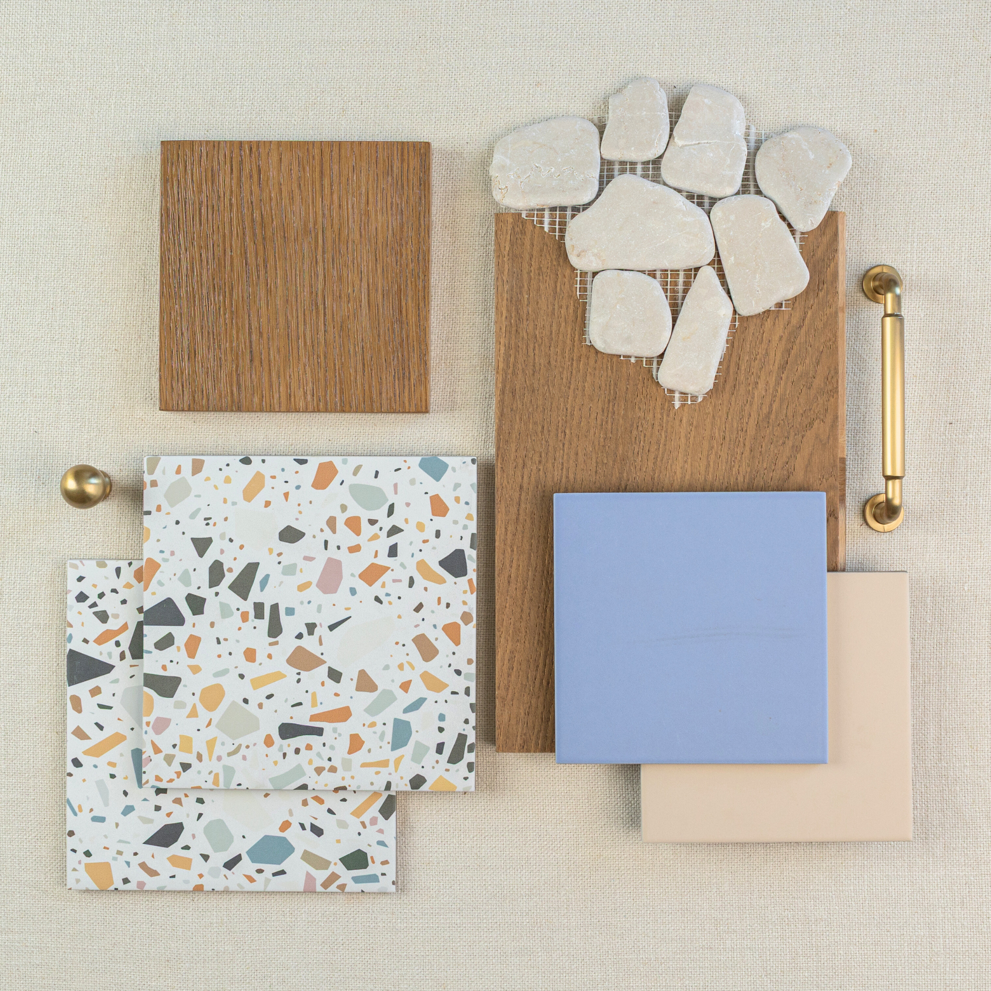 Flat lay showing wood samples, white stone mosaics, blue and beige ceramic tiles, terrazzo tiles, and brass hardware on neutral fabric background