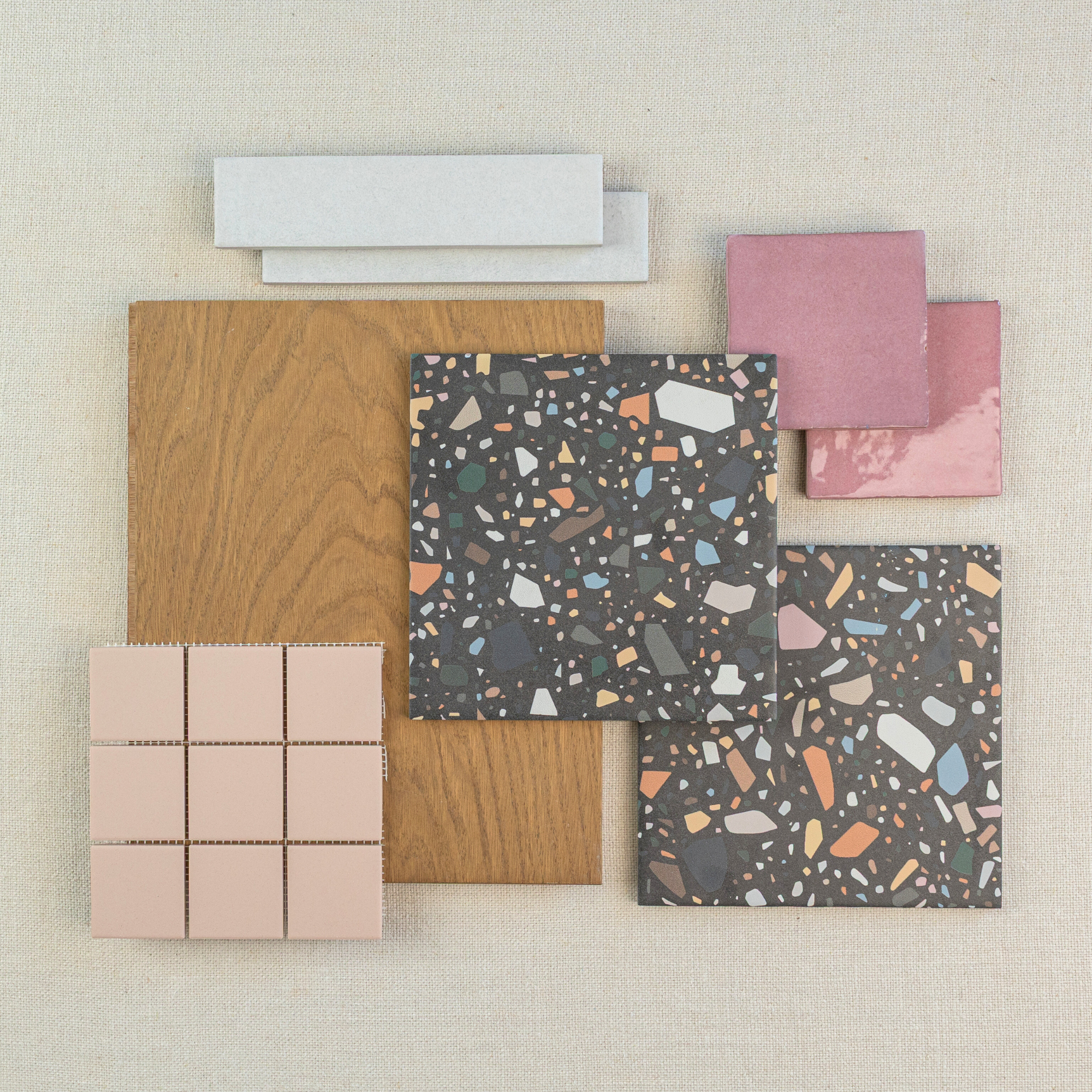 Selection of terrazzo tiles, pink ceramic squares, wood plank sample, and light gray elongated tiles on beige fabric background