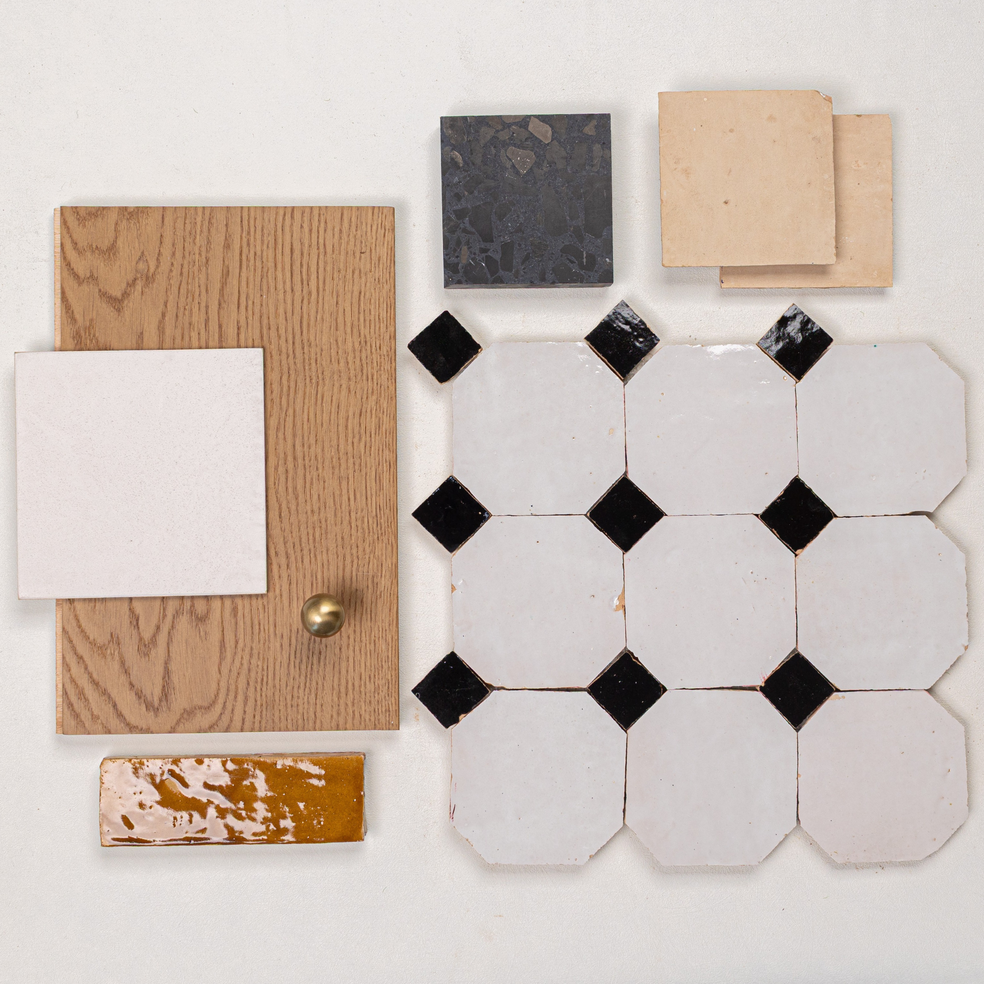 Flat lay of octagonal white tiles with black points, wood sample with brass knob, dark terrazzo, beige tiles, and glossy amber tile piece