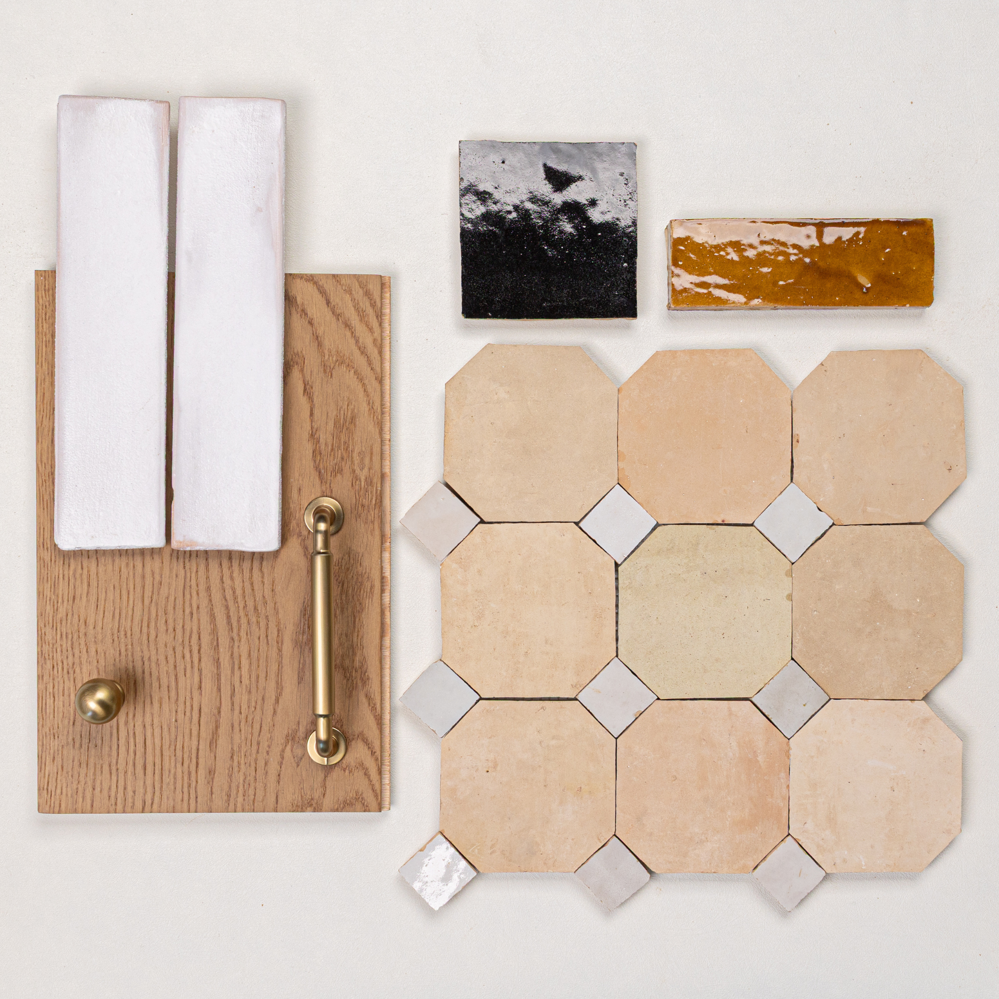 Flat lay of warm beige octagon porcelain tiles, glossy amber and black ceramic tiles, oak wood panel with brass handle and knob, and white ceramic tile samples