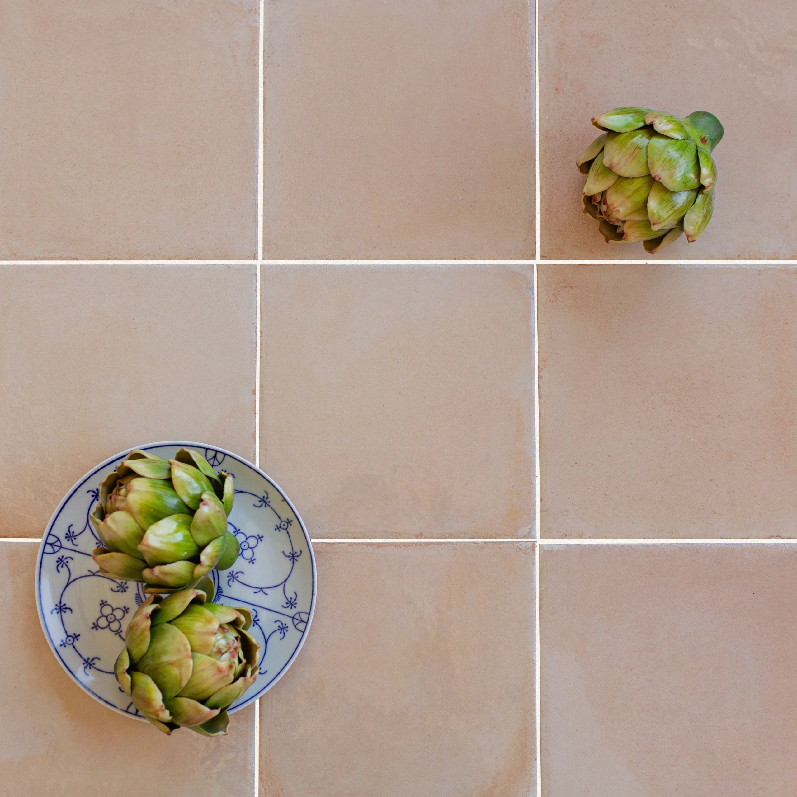 Glossy salmon-colored porcelain tiles arranged in a grid with fresh green artichokes and a patterned blue and white plate