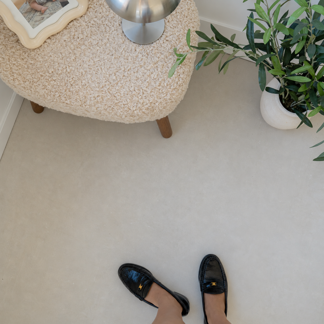 Person wearing black loafers on a light beige floor beside a boucle ottoman and potted plant