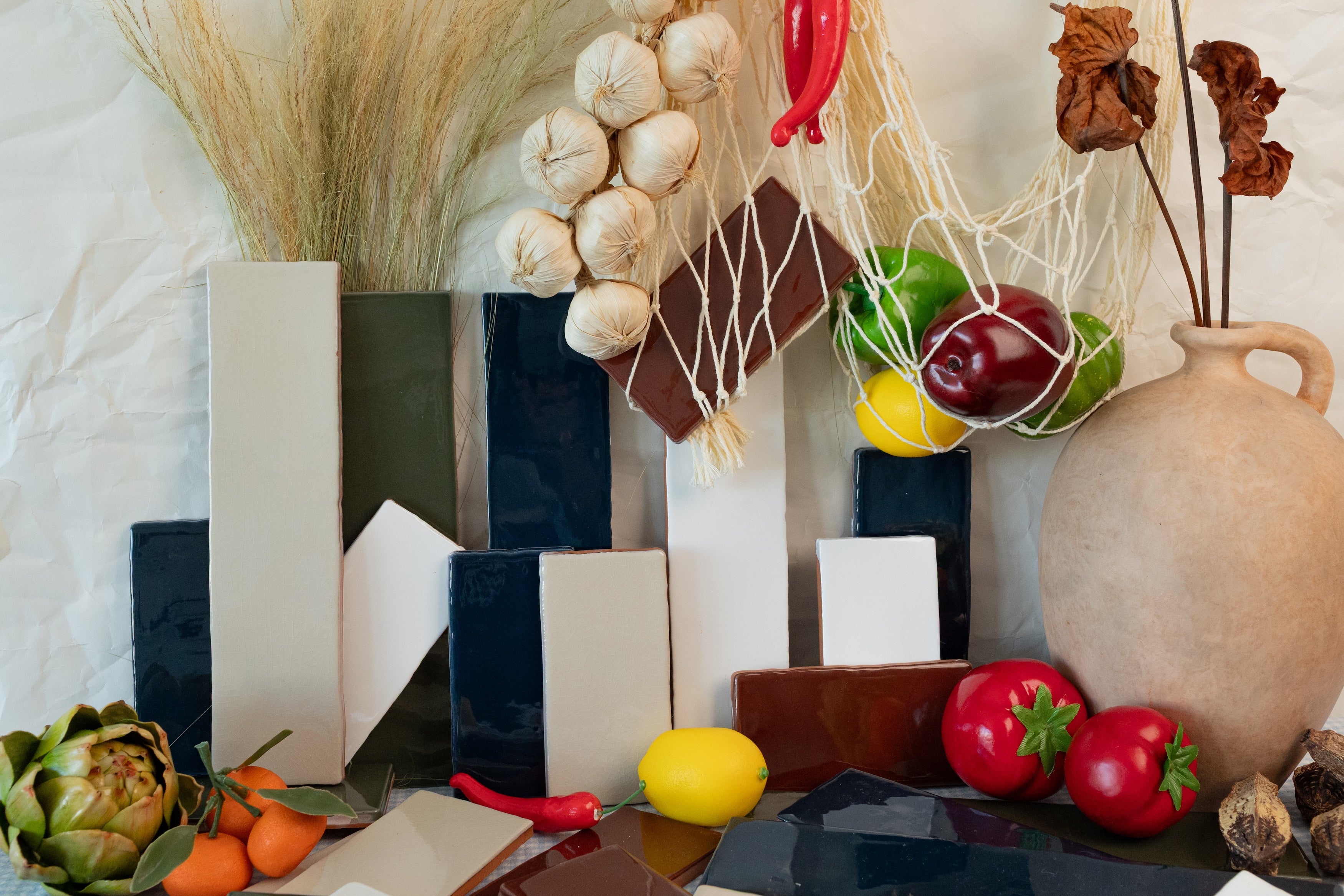 Glossy ceramic tile samples in varied blue, cream, and brown tones arranged with fresh produce and decorative dried plants