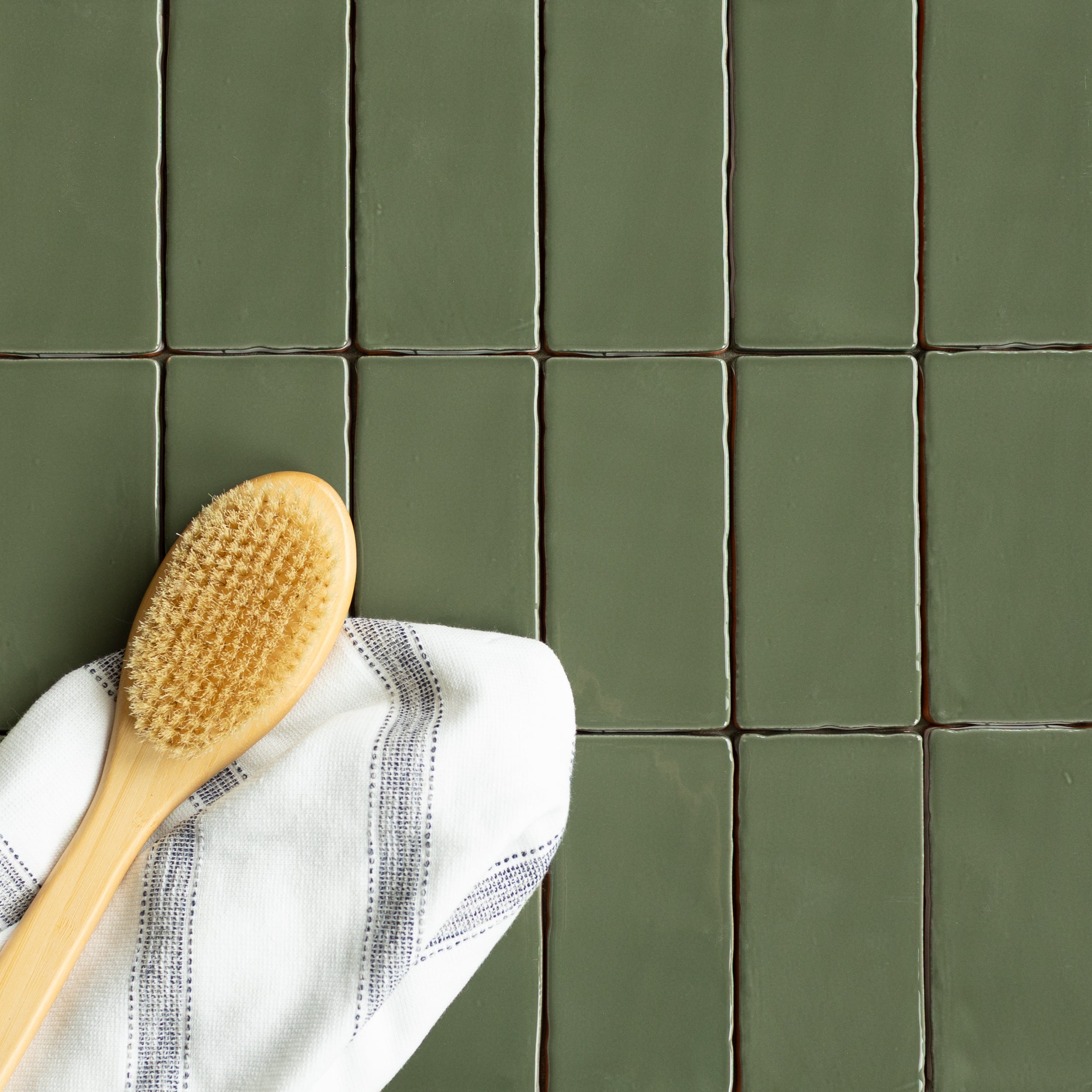 Matte moss green ceramic tiles with subtle uneven edges arranged in a clean grid alongside a wooden bath brush and striped cloth