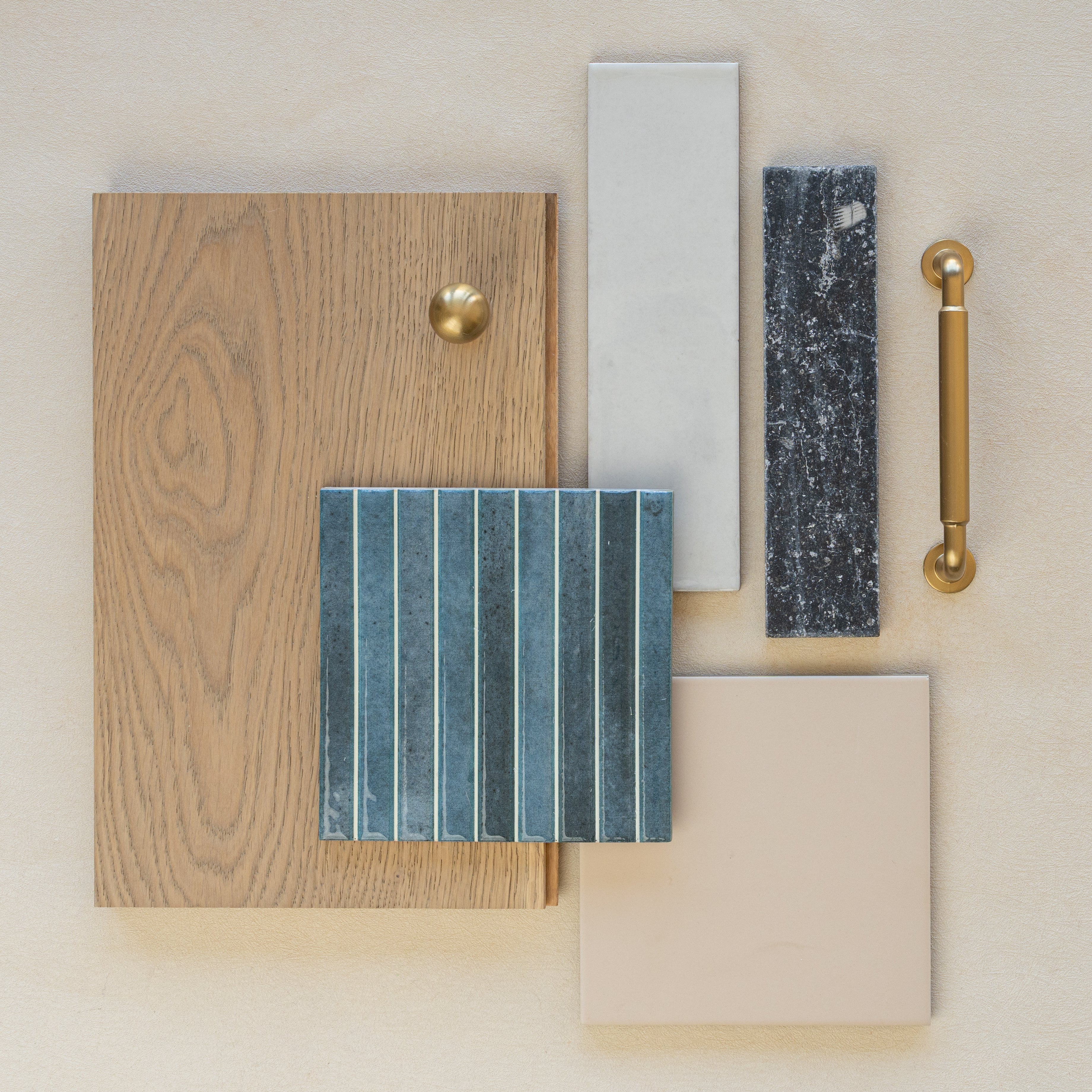 Flat lay of oak wood sample with brass hardware, beige and light gray ceramic tiles, and textured dark blue tile on neutral background