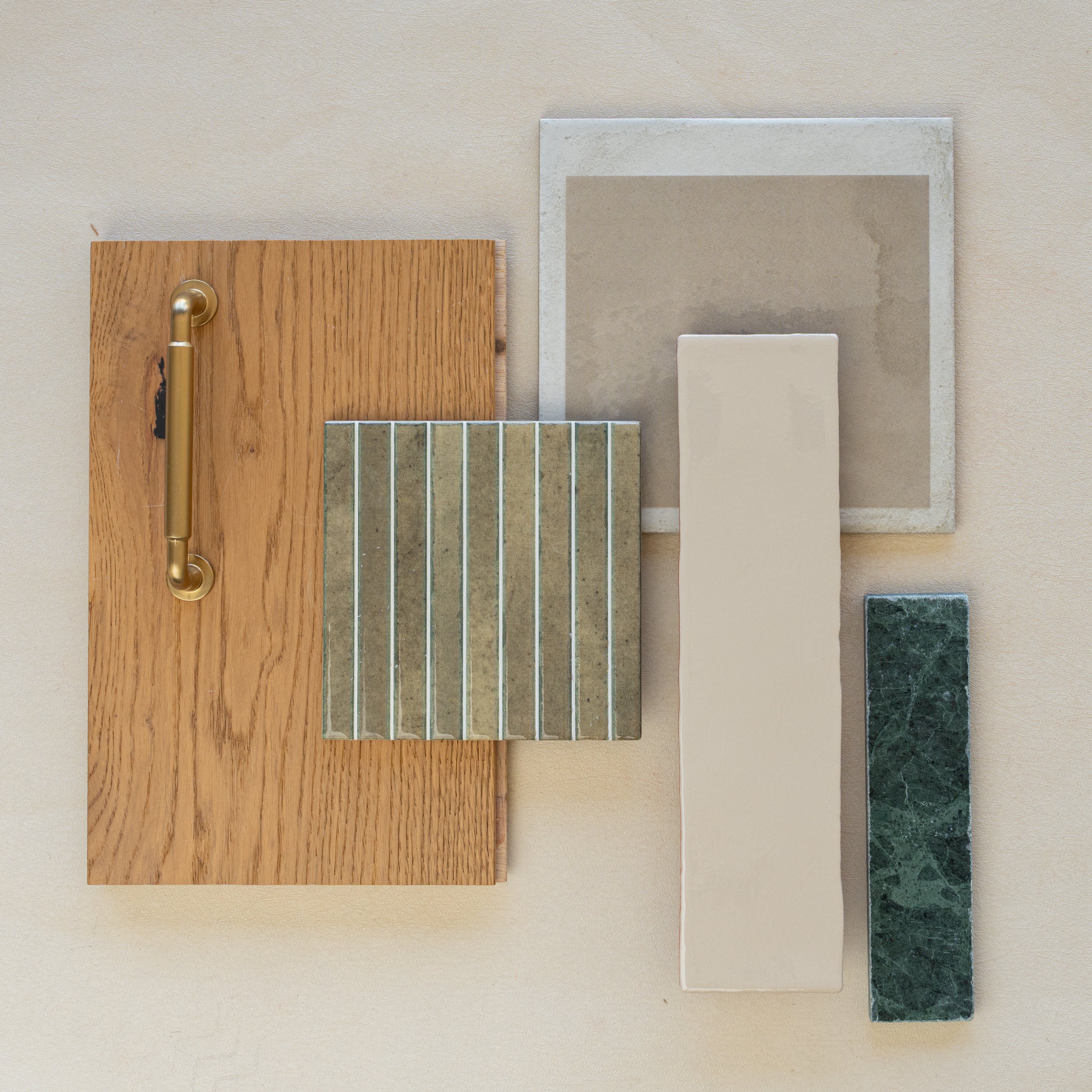 Flat lay of oak wood sample with brass handle, textured green ceramic tile, beige porcelain tile, taupe square tile, and green marble strip on neutral background