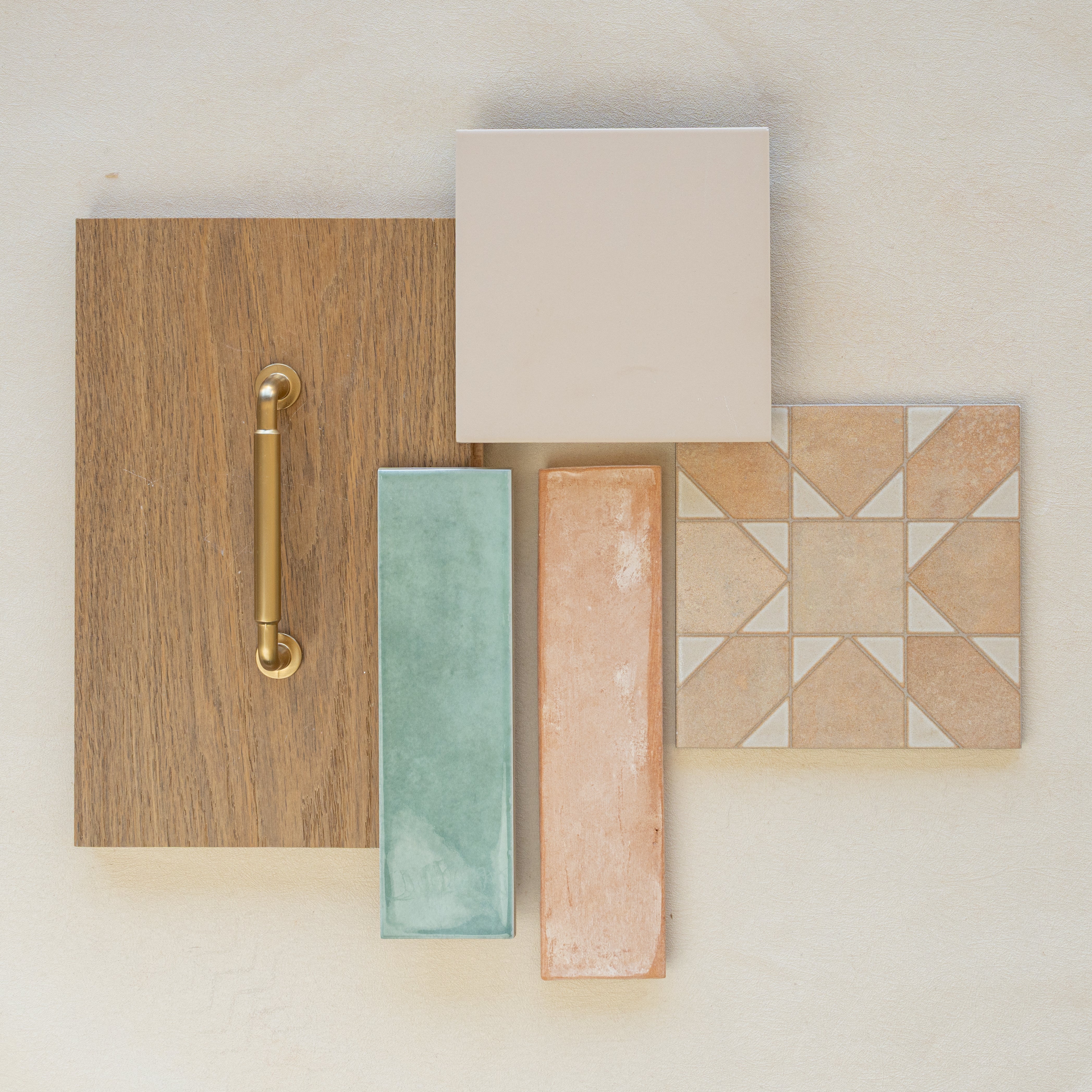 Flat lay of oak wood sample with brass handle, beige and patterned ceramic tiles, green glossy tile, and terracotta tile on neutral background
