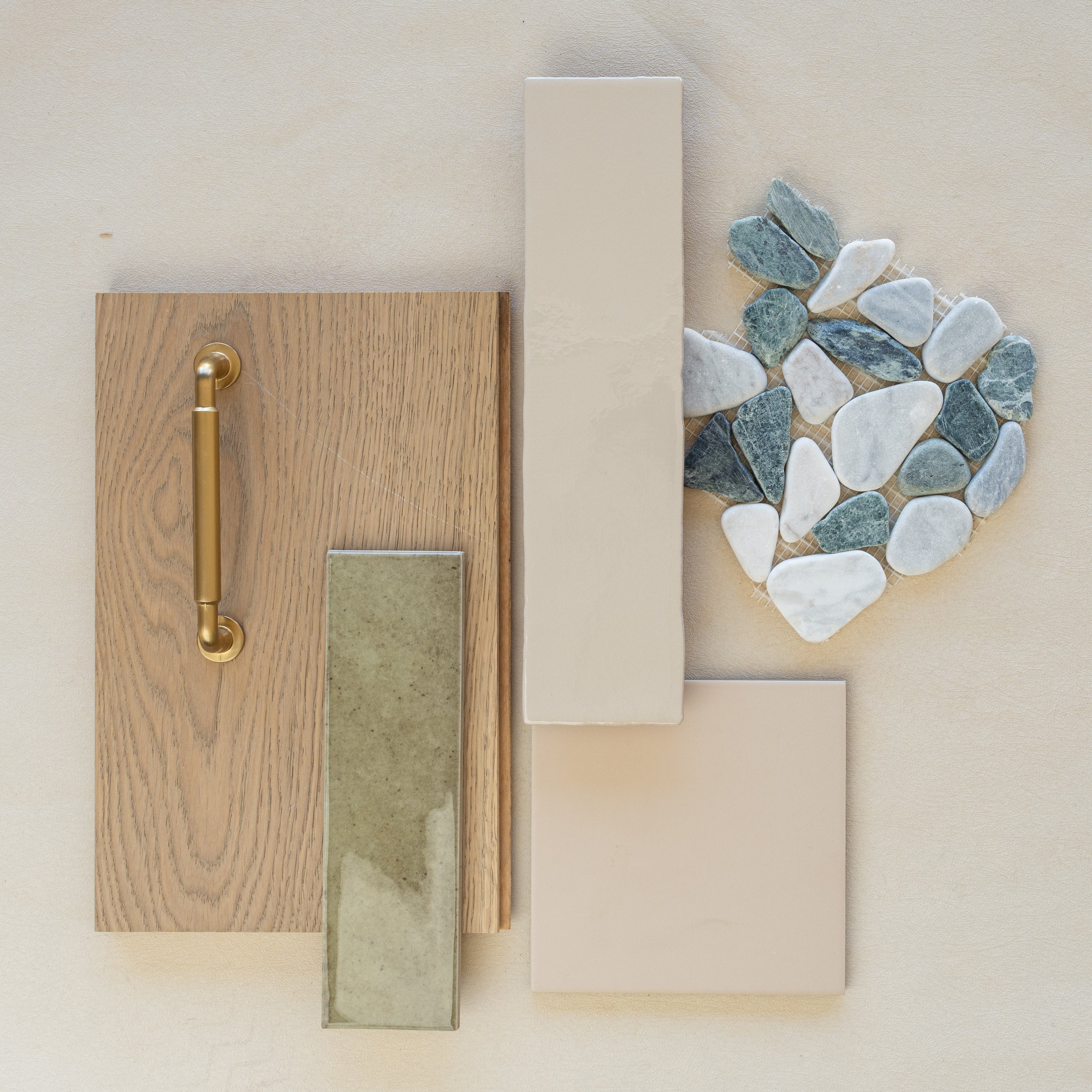 Flat lay of oak wood sample with brass handle, olive green ceramic tile, beige porcelain tiles, and natural stone pebble mosaic