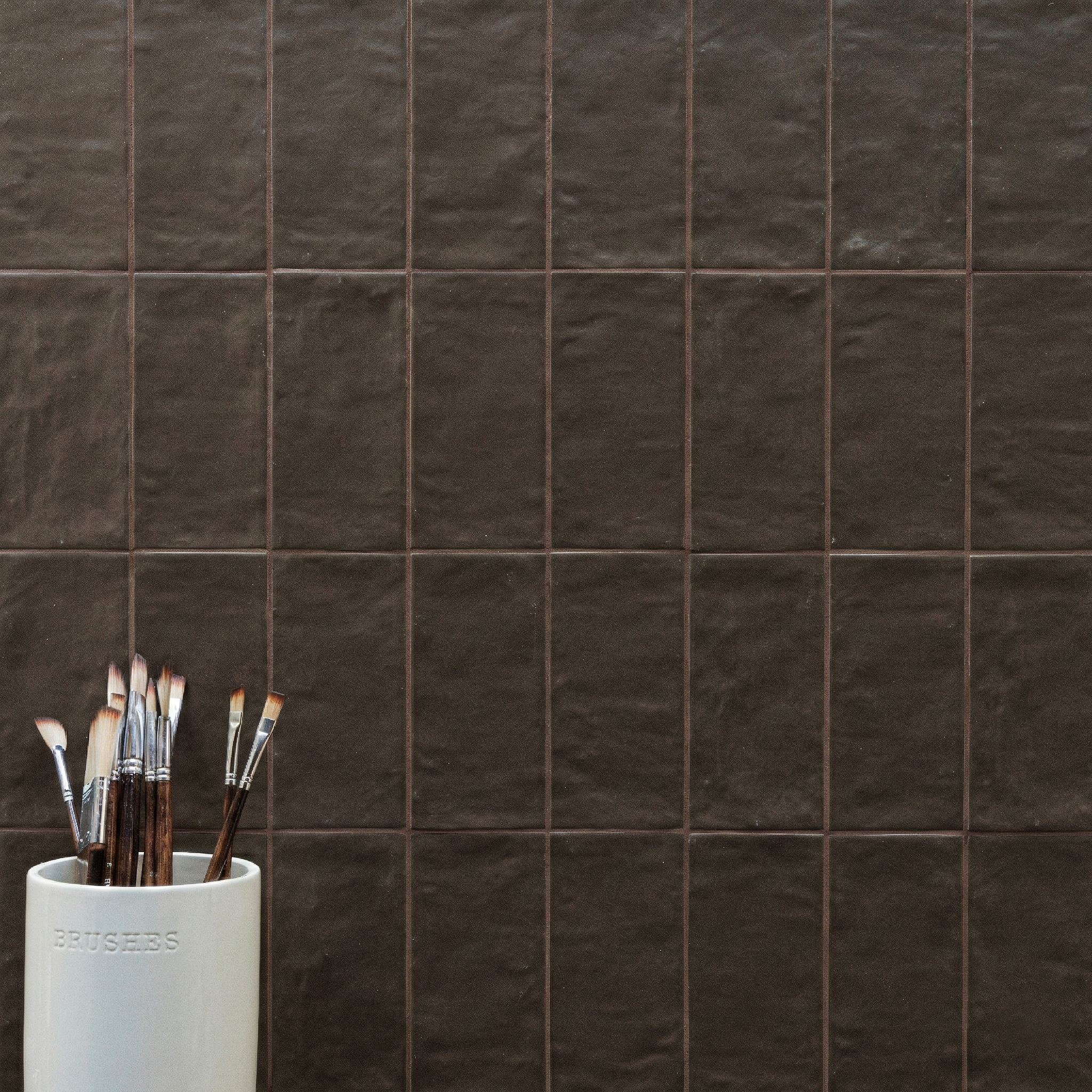 Square dark brown textured ceramic tiles arranged in a grid with a white container holding various paintbrushes at the corner