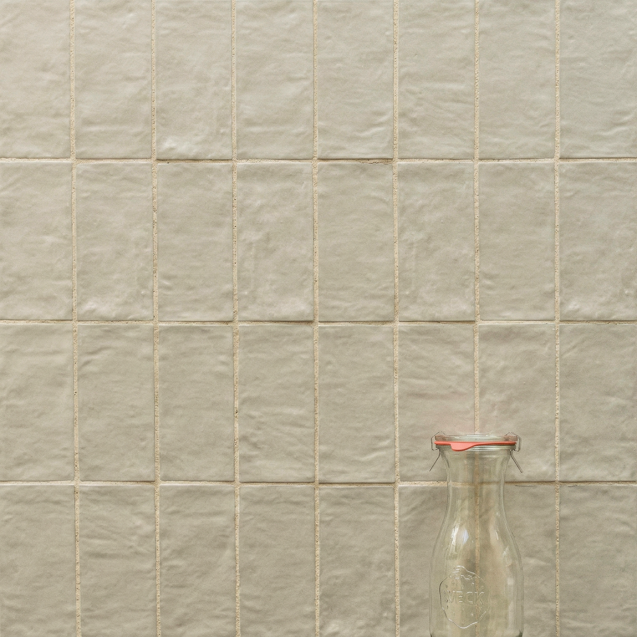 Square warm beige ceramic tiles with a textured surface arranged in a grid behind a clear glass bottle with a red lid