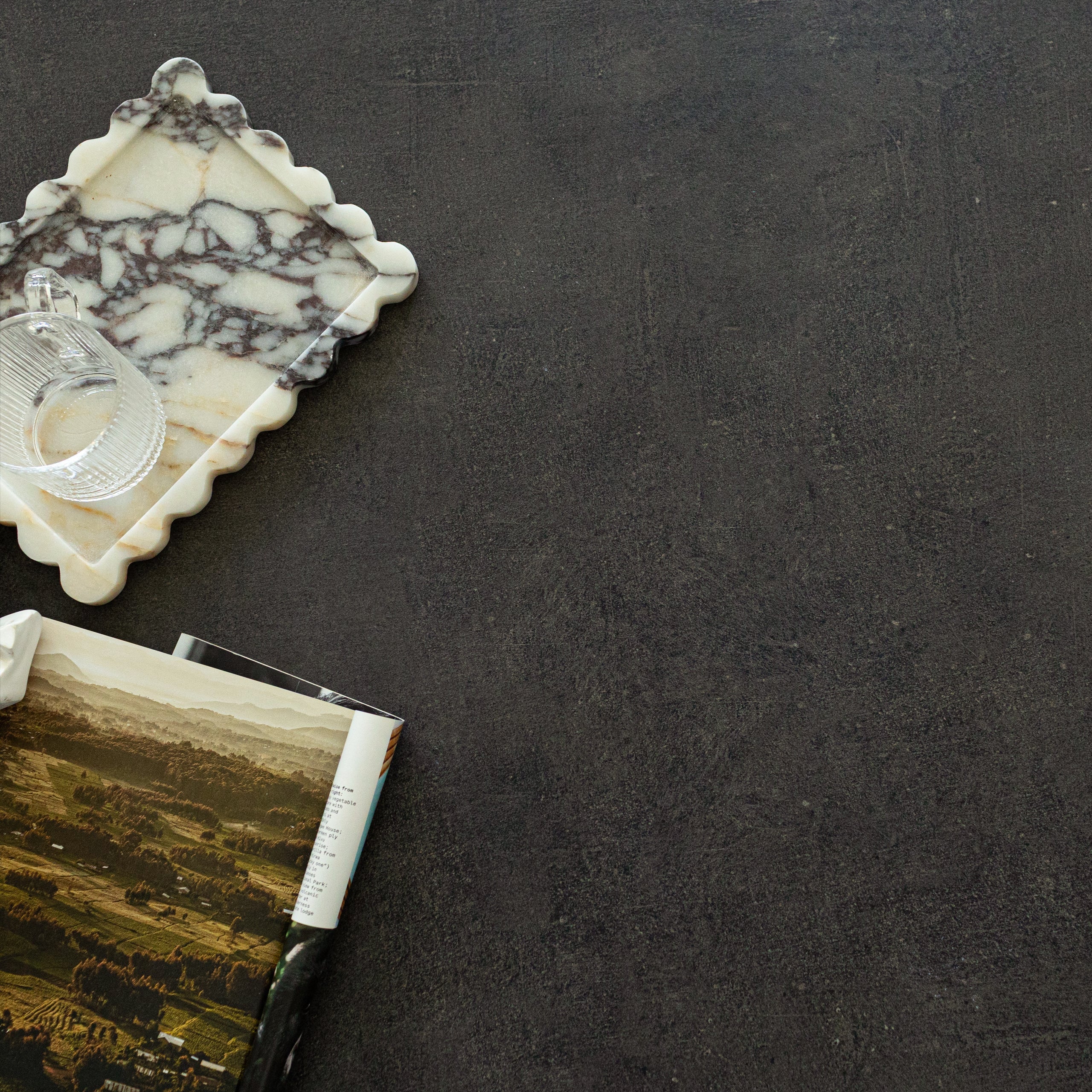 Dark slate-effect porcelain tile surface with marble tray holding a glass cup and a partially rolled landscape magazine nearby
