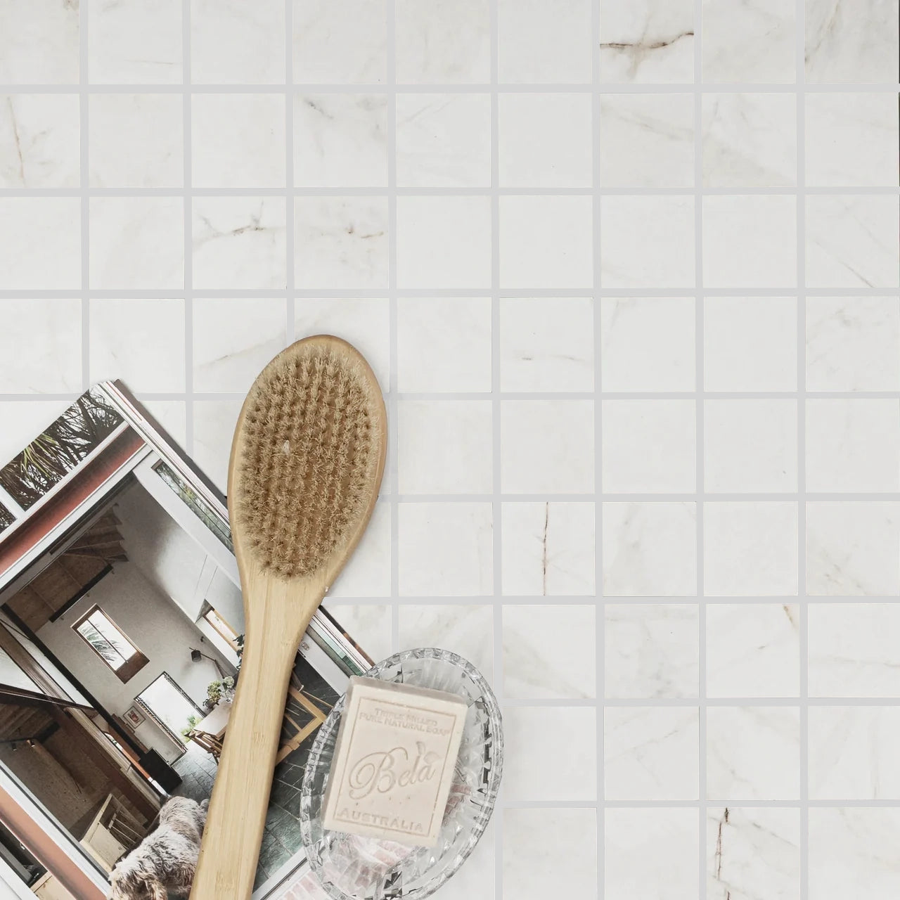 Flat lay showing white marble-look porcelain mosaic tile with wooden brush, soap bar, and open magazine on surface