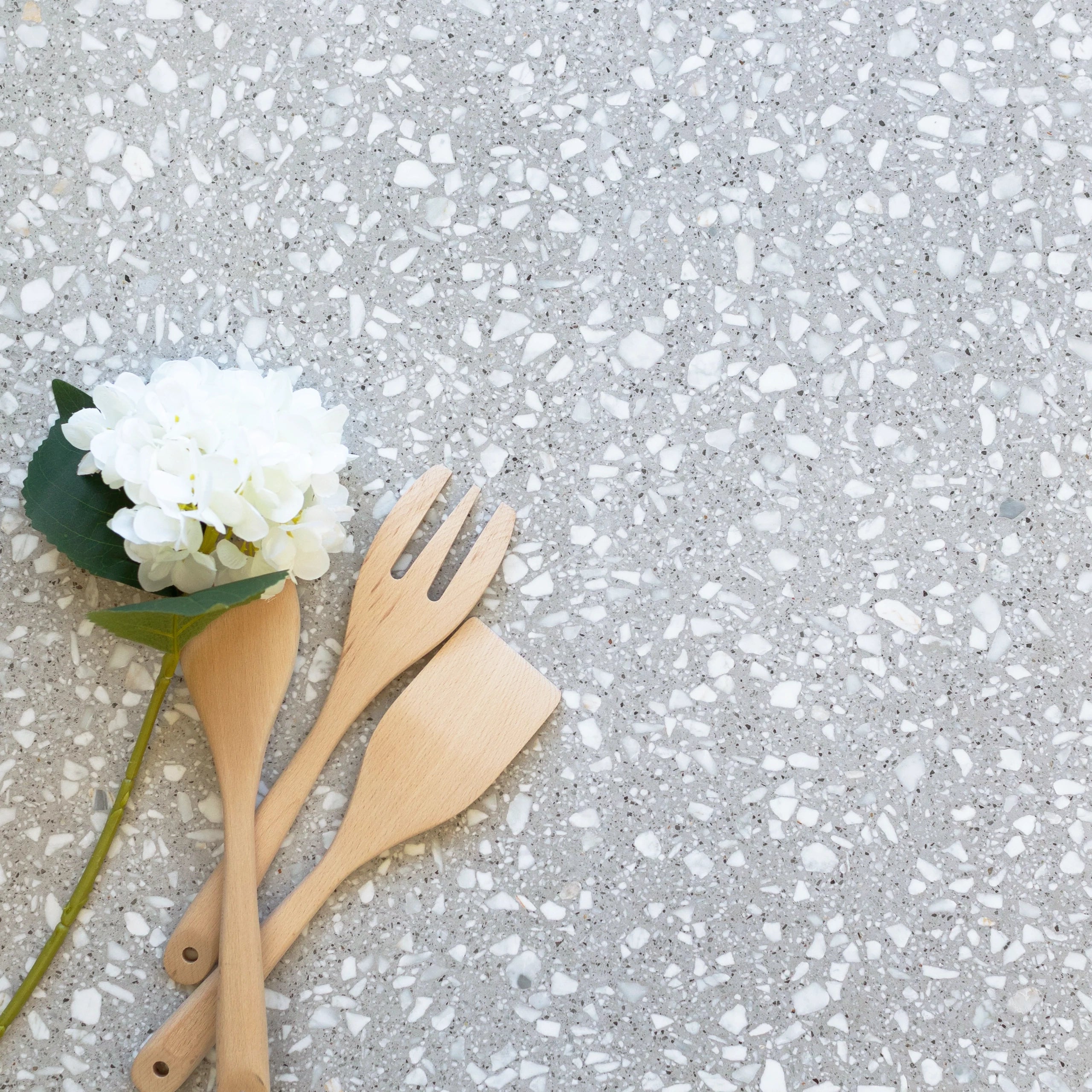 Light gray terrazzo surface with white chips, arranged with a white flower and three wooden kitchen utensils nearby