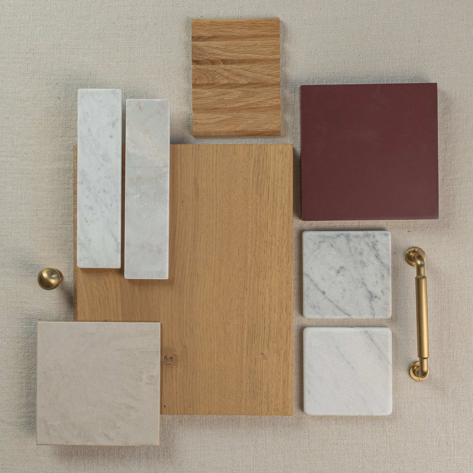 Flat lay of oak wood samples, marble tiles, a burgundy ceramic tile, beige stone tile, and brass hardware on neutral fabric
