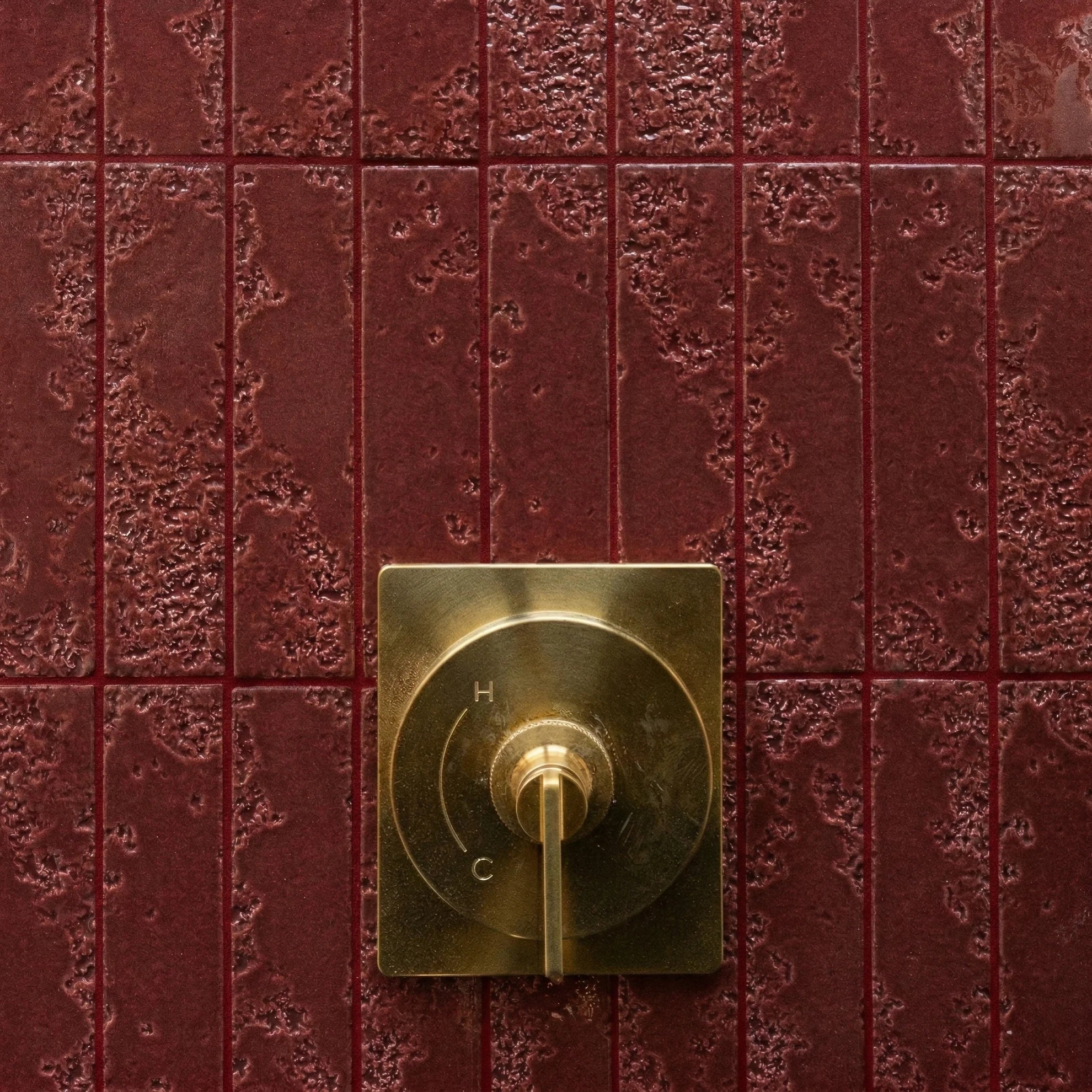 Close-up of a textured deep red ceramic tiled wall with a brushed brass temperature control knob marked H and C.