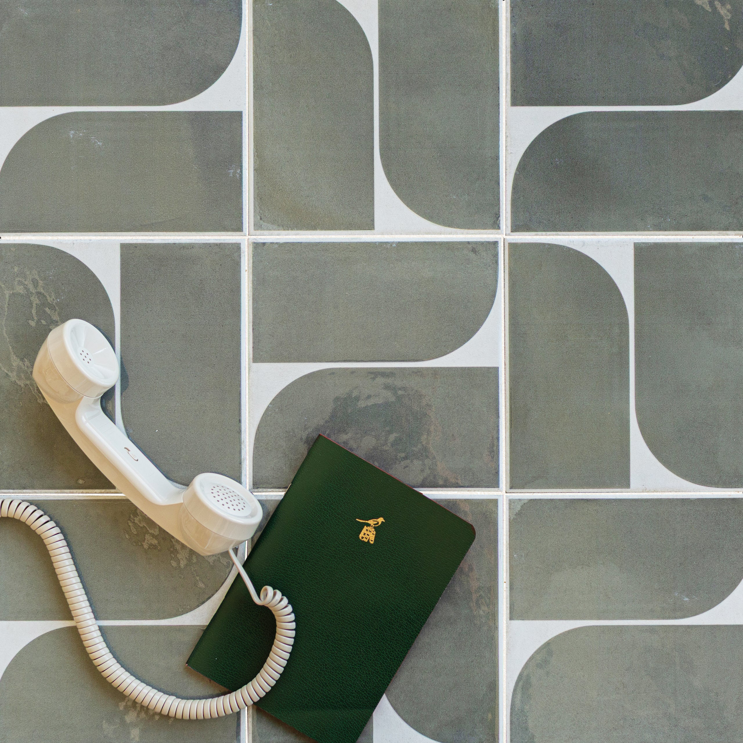 Matte porcelain tiles in muted gray-green tones with curved geometric patterns beside a cream telephone handset and dark green notebook