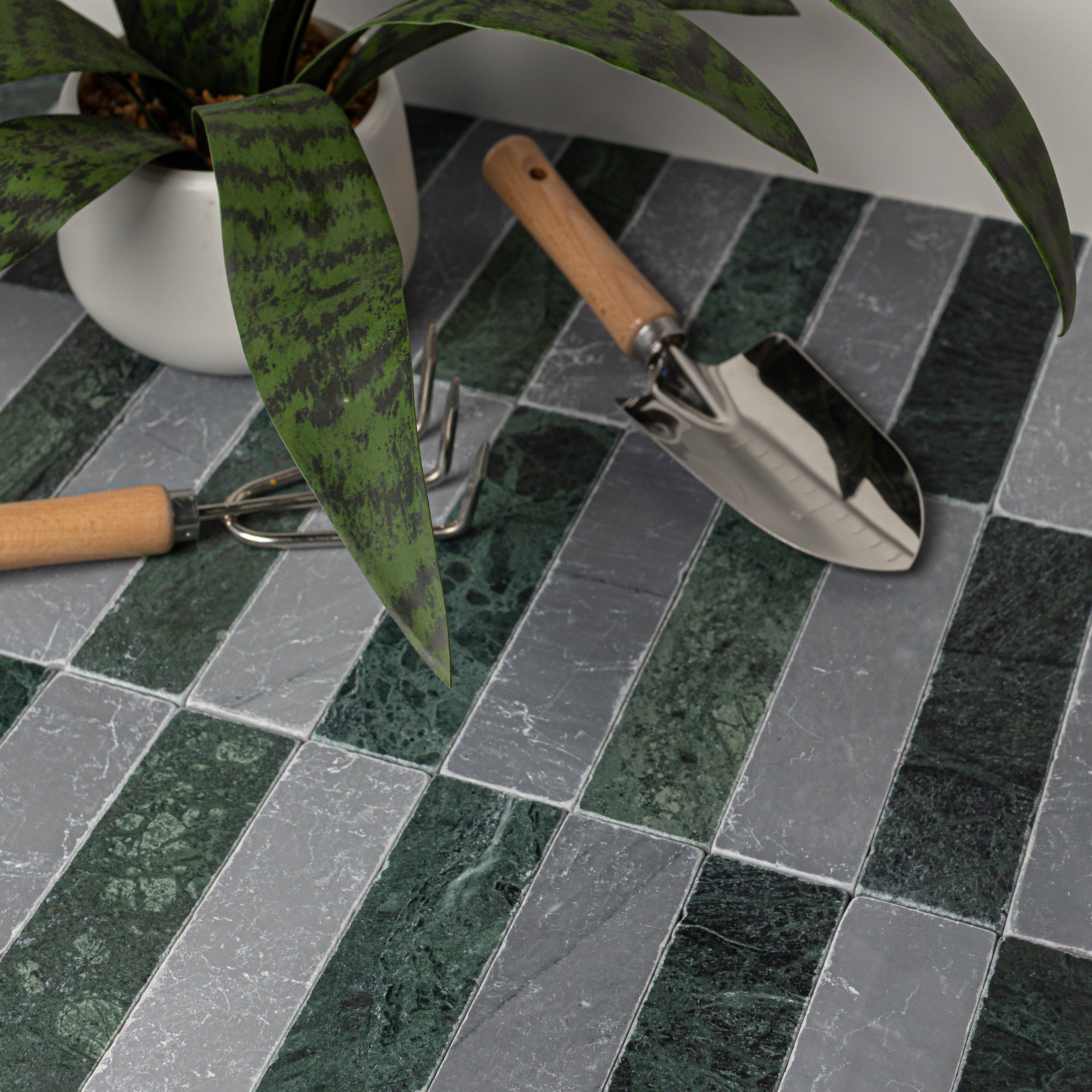 Rectangular green and gray tumbled stone tiles arranged in a staggered pattern with gardening tools and a leafy plant nearby