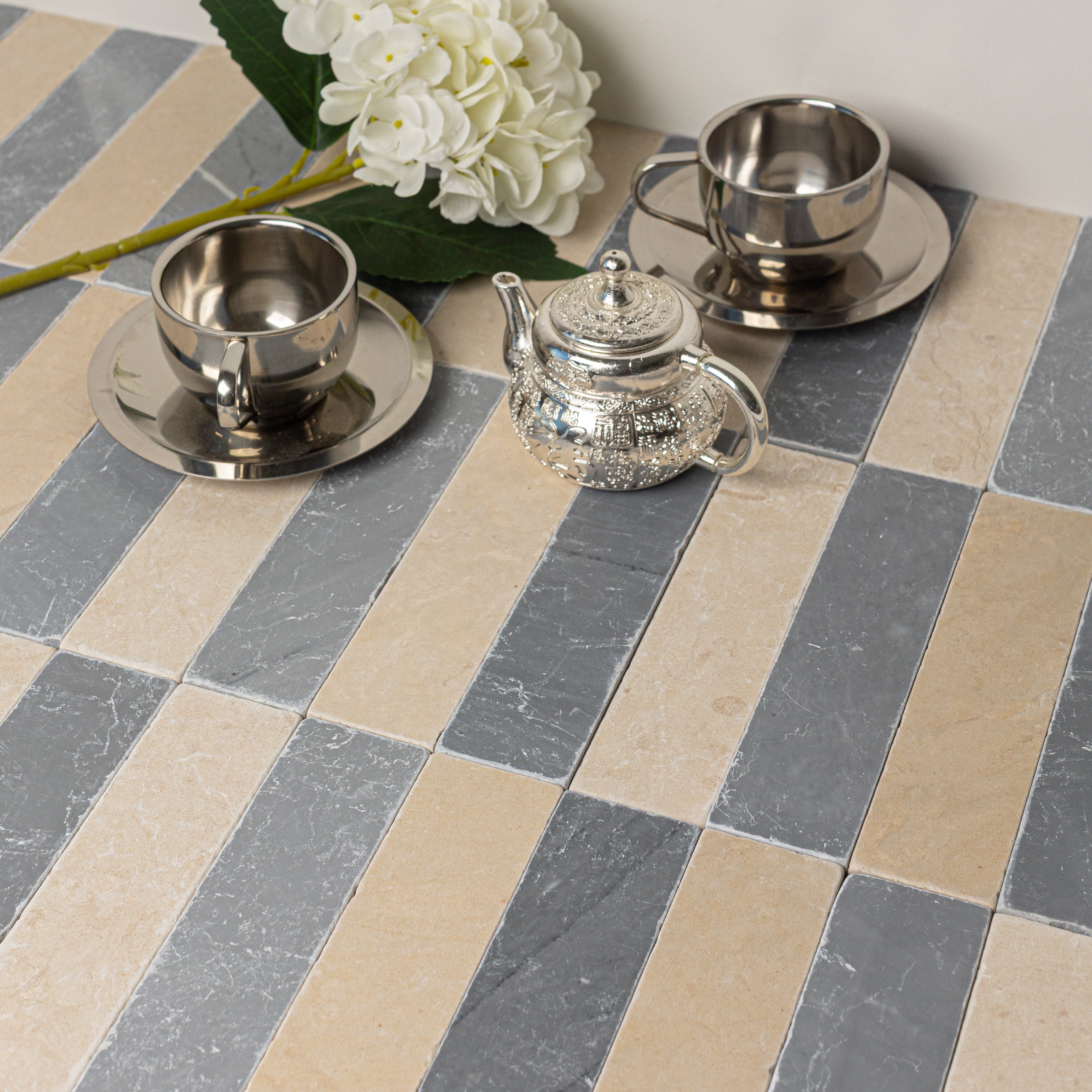 Stainless steel tea set with cups and saucers arranged on alternating beige and gray rectangular tiles with white flower accents