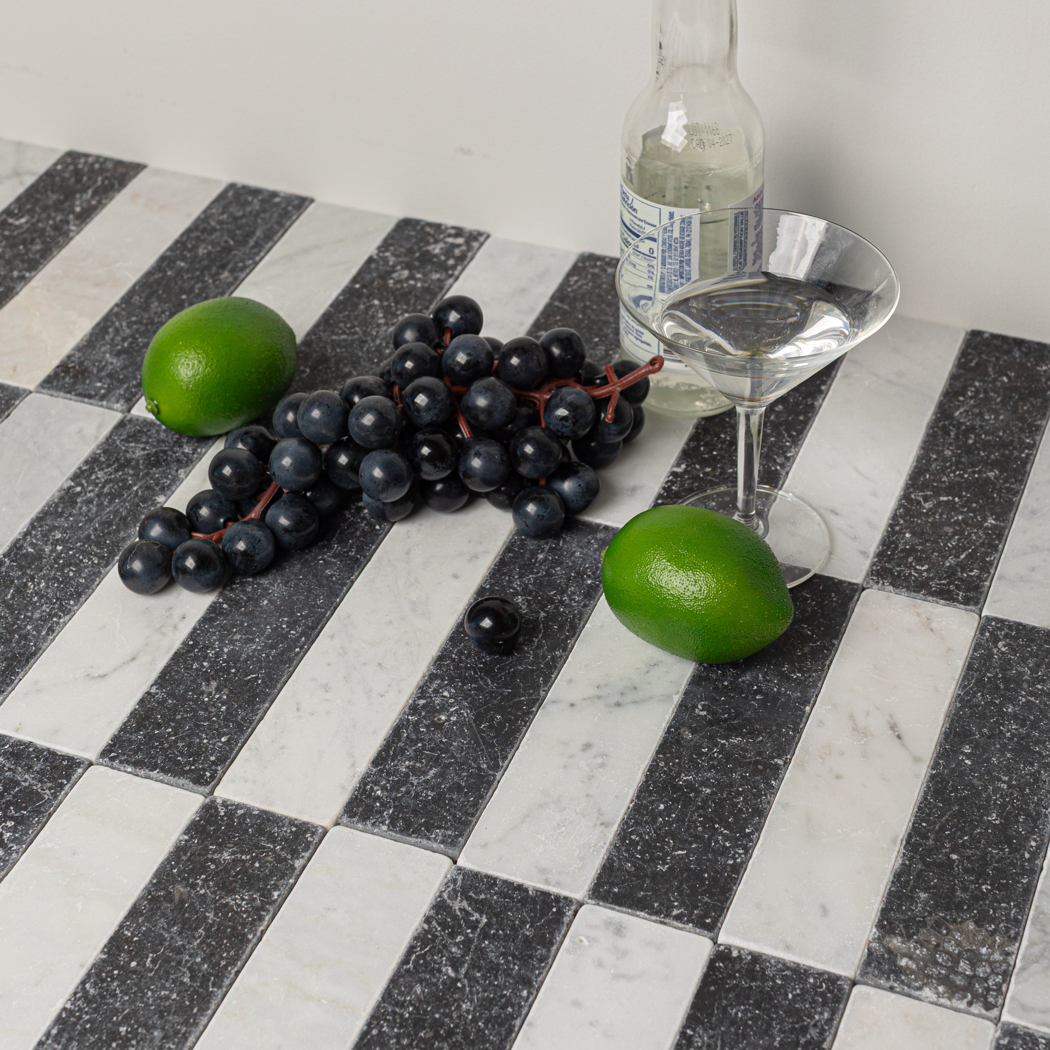 Alternating black and white rectangular stone tiles with natural texture holding a bottle, cocktail glass, grapes, and limes