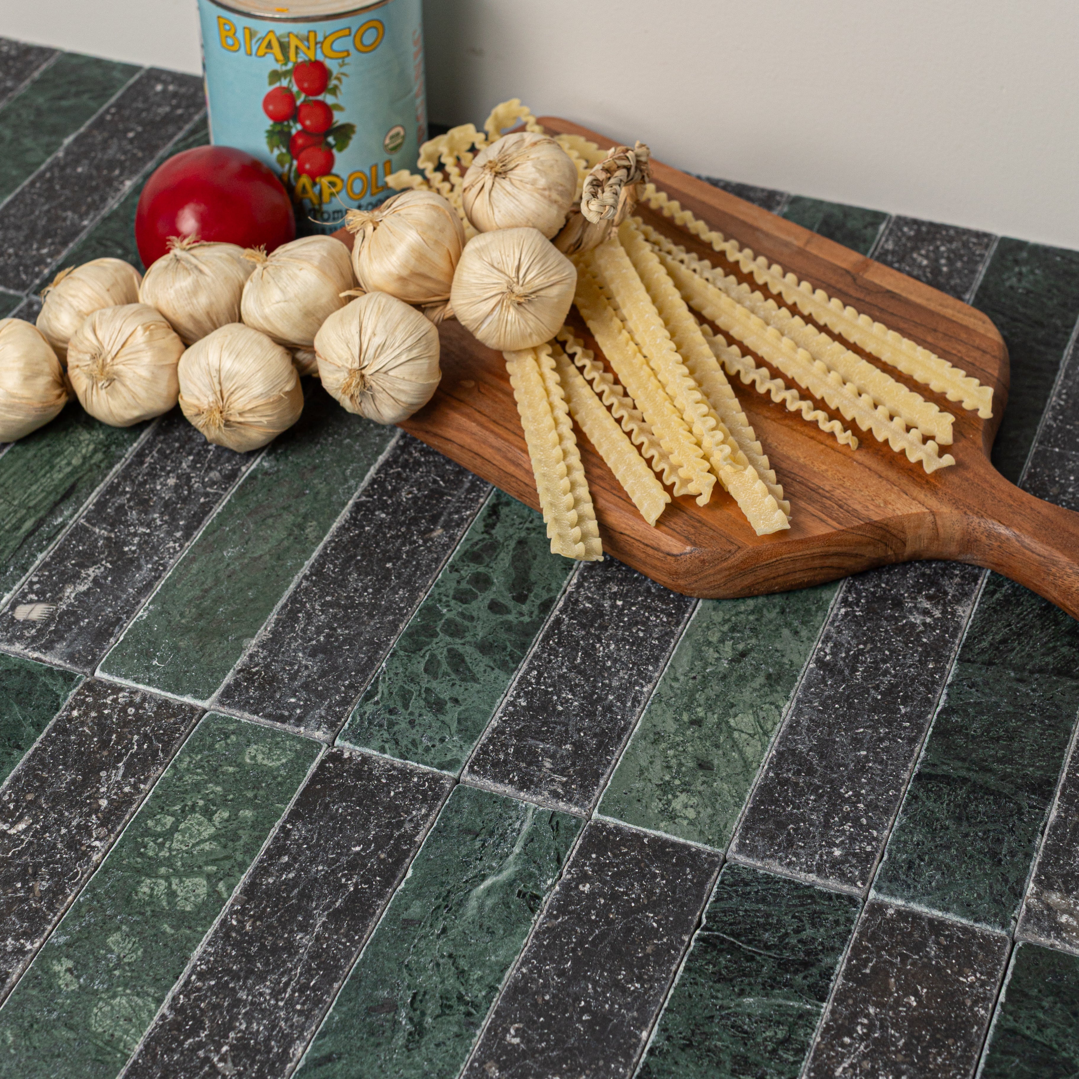 Dark green and black textured stone tiles arranged in alternating stripes with garlic bulbs, pasta, tomato, and a vintage tin on top