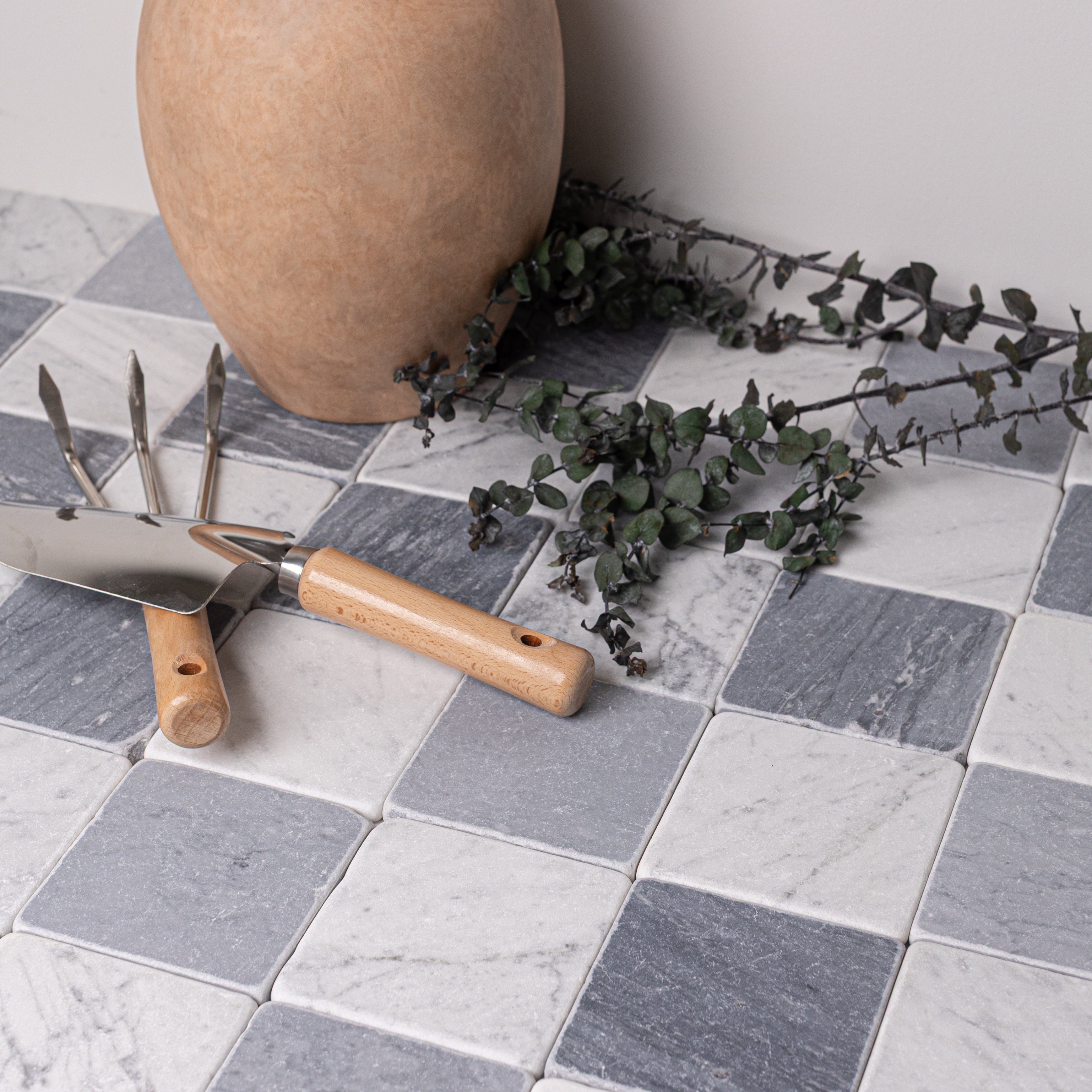 Checkerboard pattern of gray and white tumbled stone tiles with gardening tools, dried eucalyptus, and a textured clay pot nearby