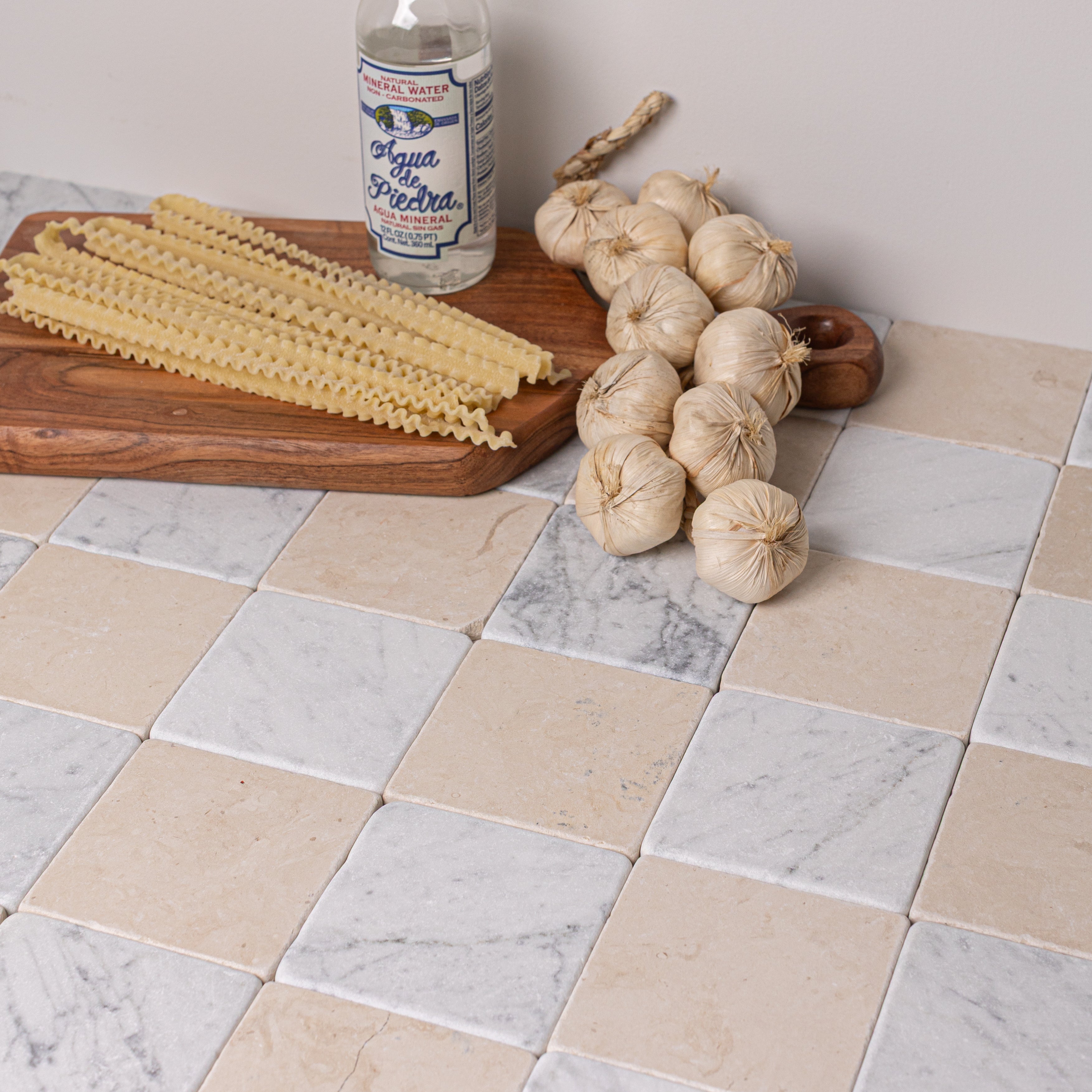 Close-up of beige and gray marble tiles with wood cutting board, garlic bundle, uncooked pasta, and mineral water bottle arranged on top
