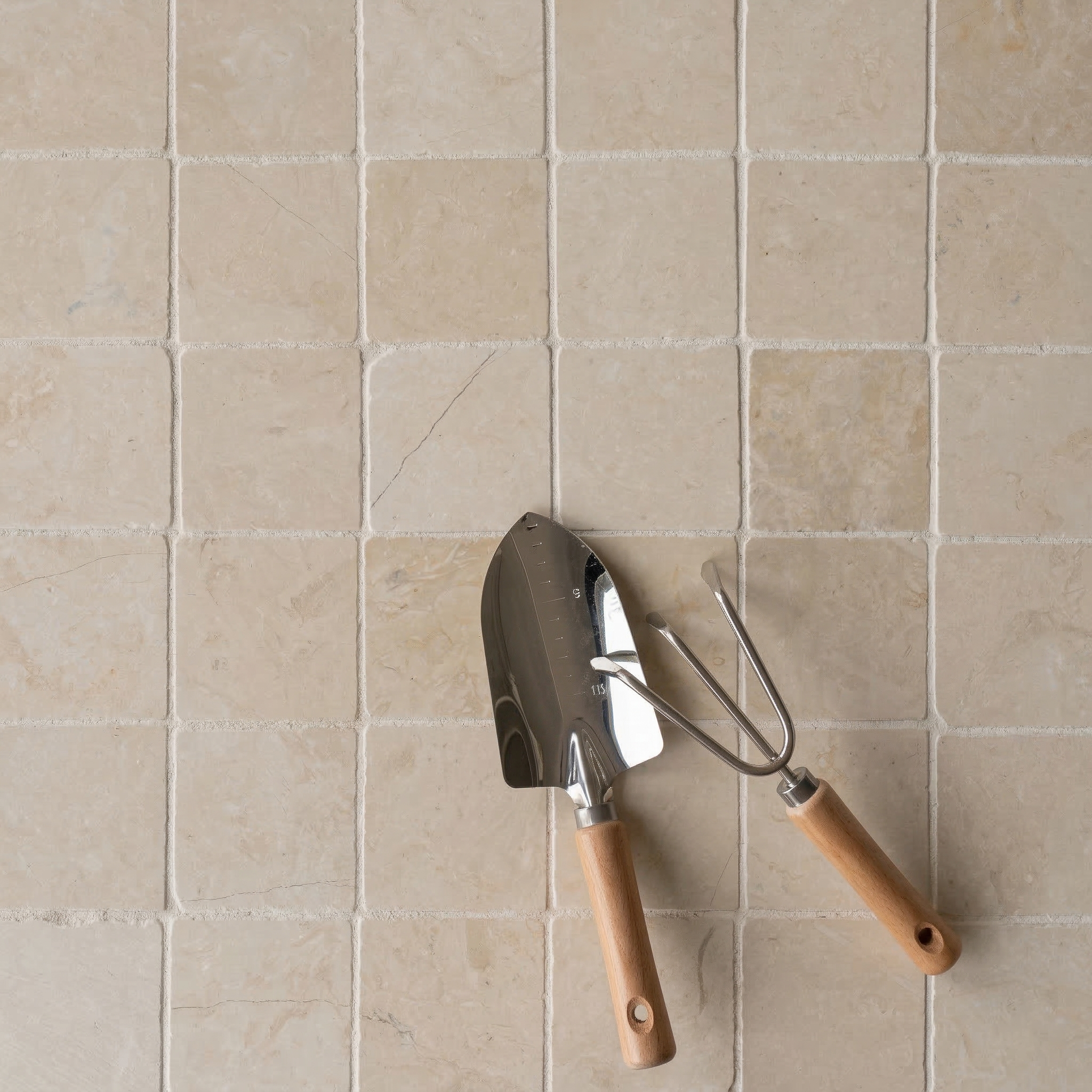 Square tumbled stone tiles in soft beige tones with natural texture, paired with wooden-handled garden tools on surface