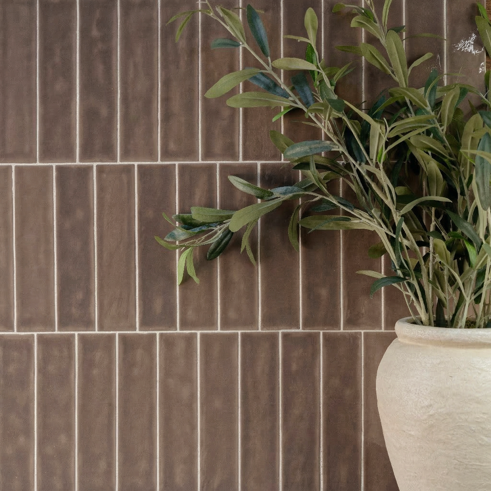 Glossy umber ceramic tiles with a subtle variegated finish arranged vertically behind a beige clay pot holding green leafy branches
