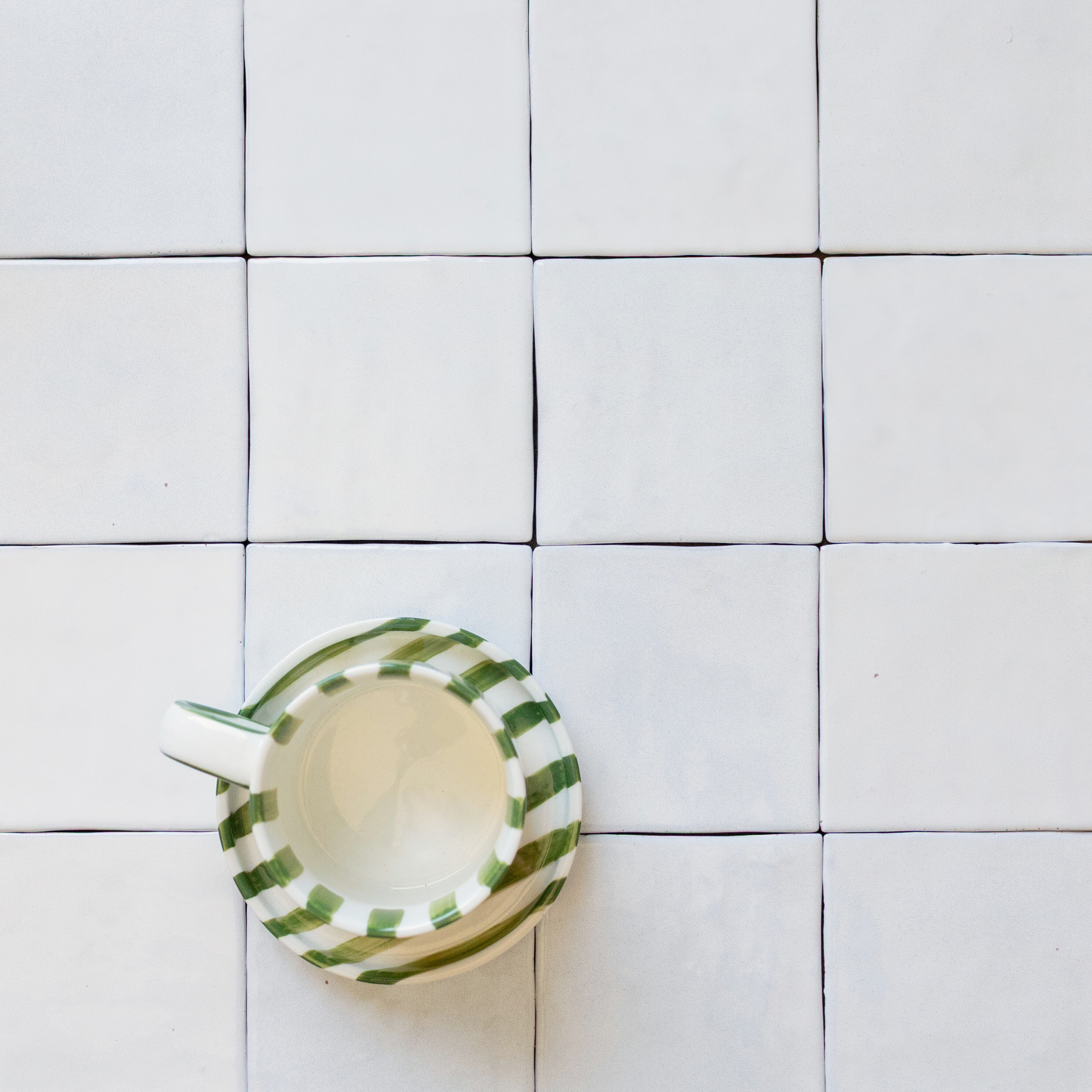 Matte white square ceramic tiles arranged in a grid with a green-striped ceramic cup placed near the bottom left corner