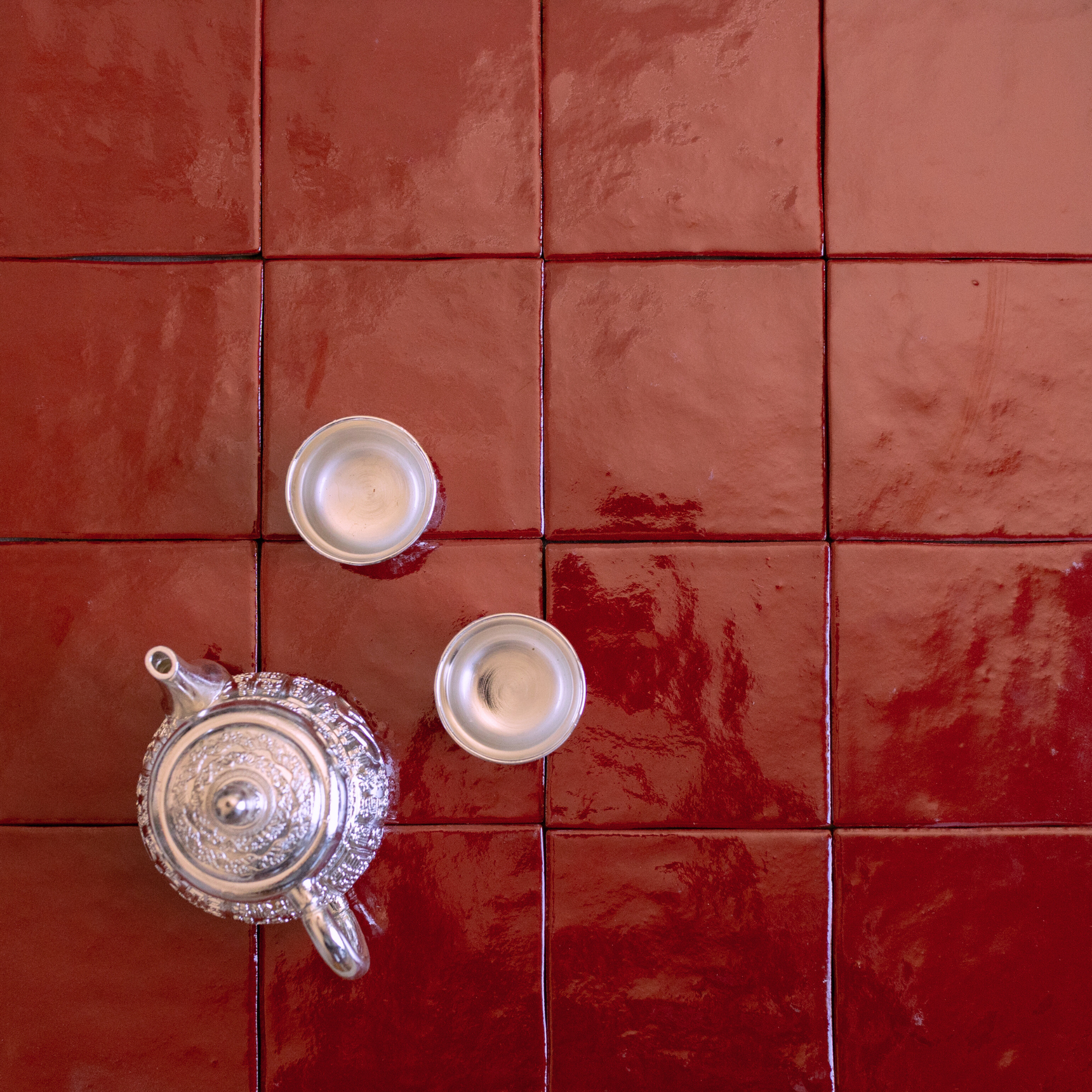 Glossy deep red square ceramic tiles with an ornate silver teapot and two small silver cups placed on top