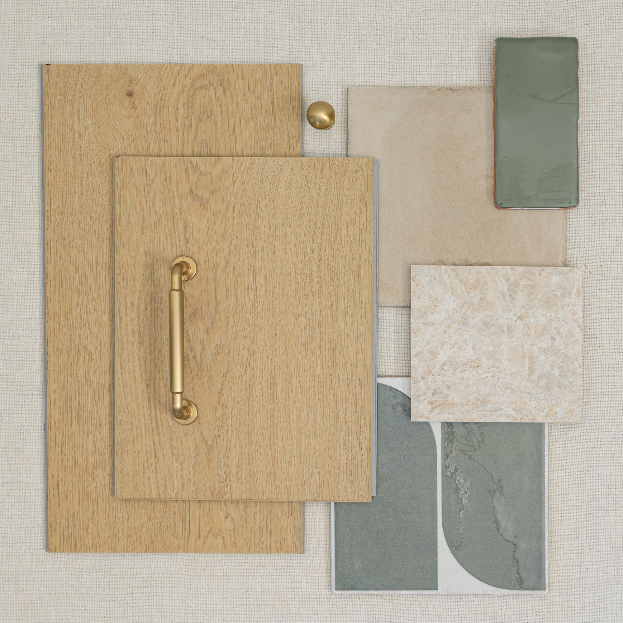 Flat lay of light oak wood cabinet fronts with brass handle and knob, beige, green, and marble tile samples on fabric background