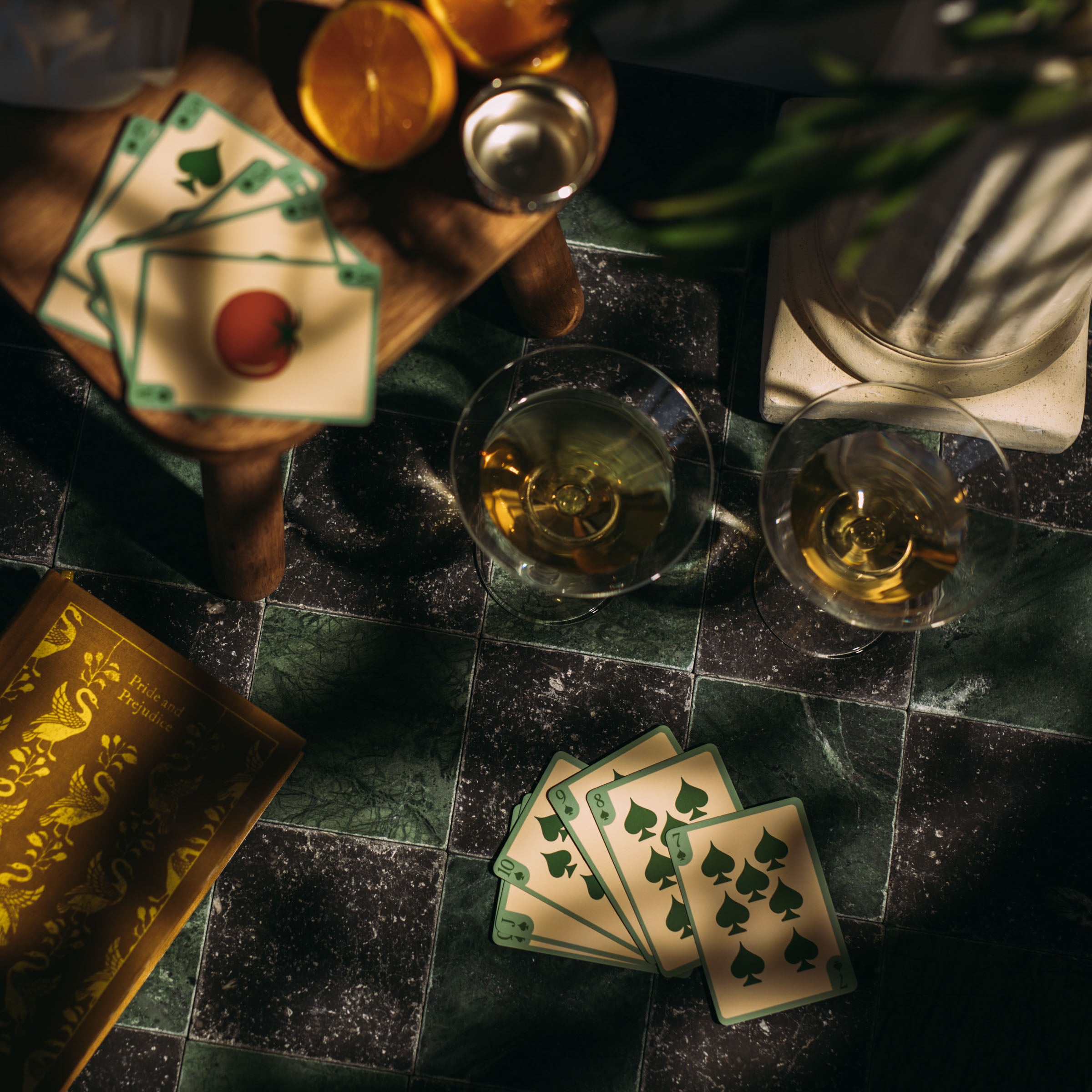 Overhead view of dark green and black marble tiles with playing cards, whiskey glasses, and a wooden cutting board with oranges