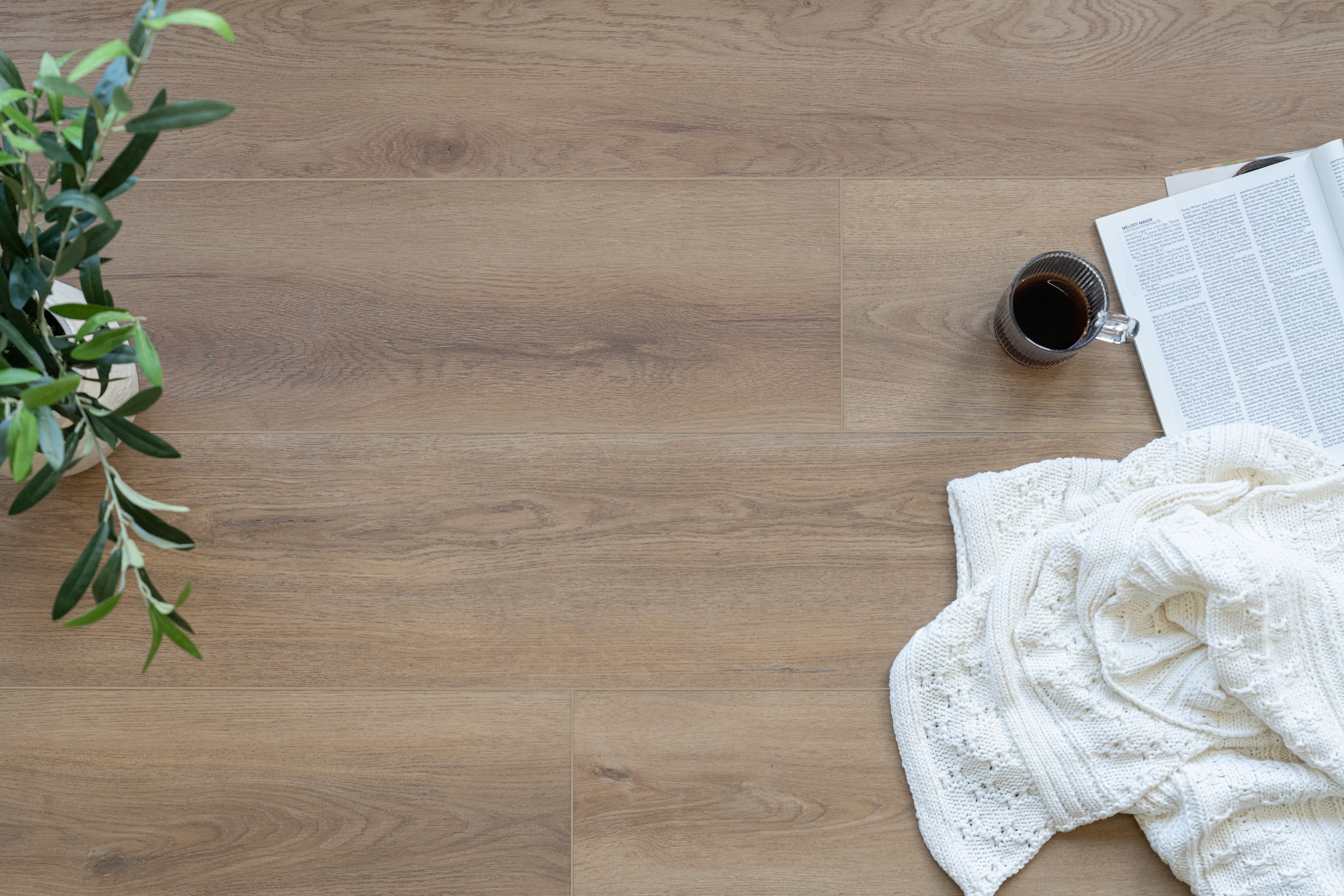 Warm-toned vinyl flooring with subtle wood grain, next to a leafy plant, a cozy white knit blanket, a book, and a cup of coffee.