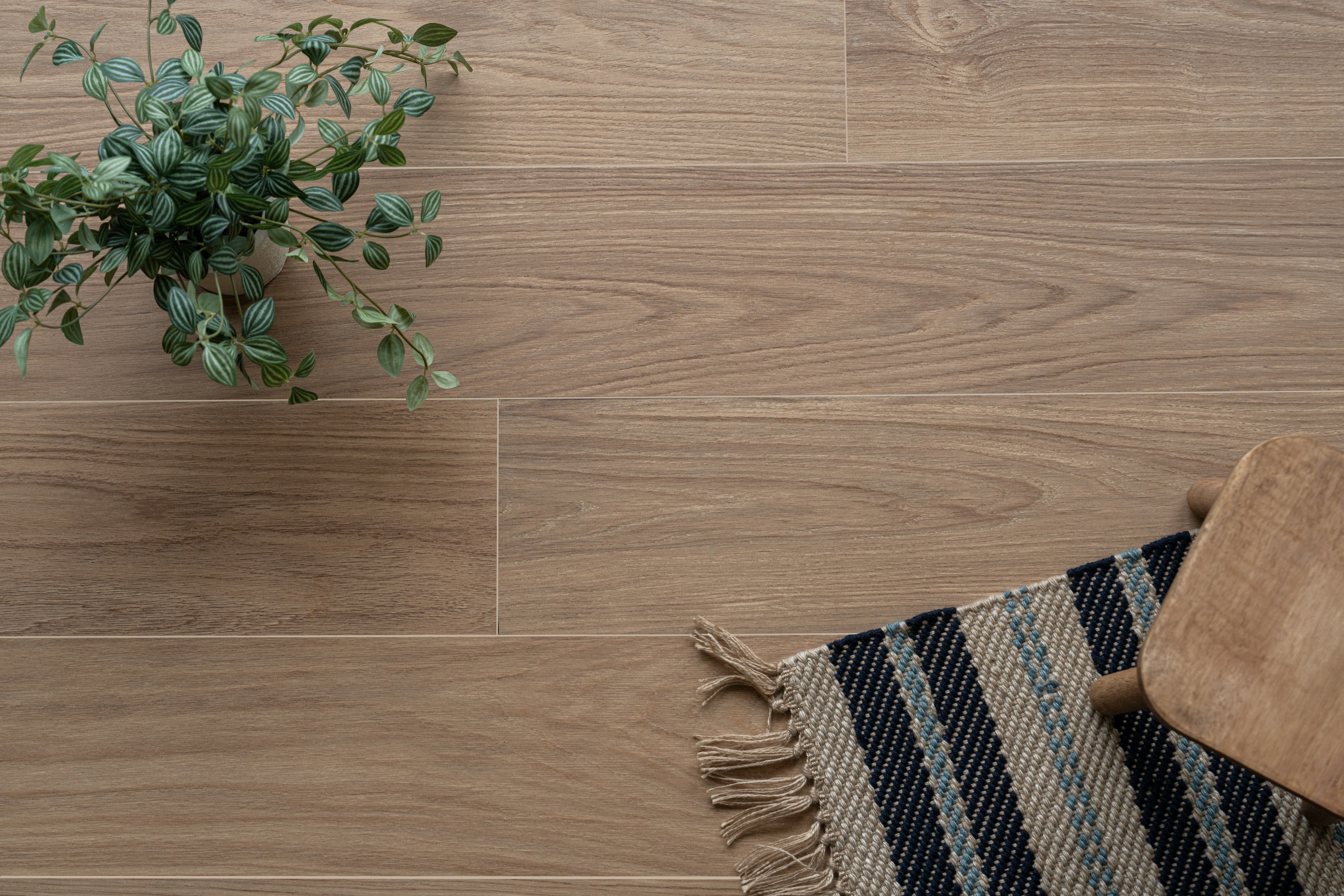 Light brown engineered wood flooring with natural grain, accompanied by a leafy plant and a woven rug with a wooden stool leg visible