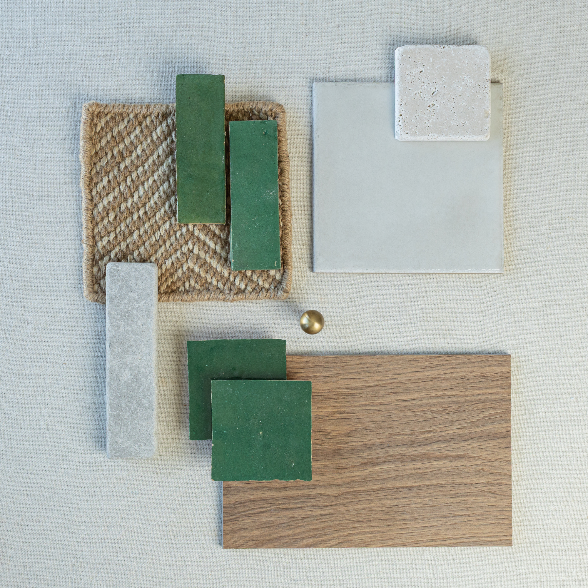 Flat lay of glossy green ceramic tiles, light wood sample, textured woven fabric, gray stone tile, and brass knob on pale background