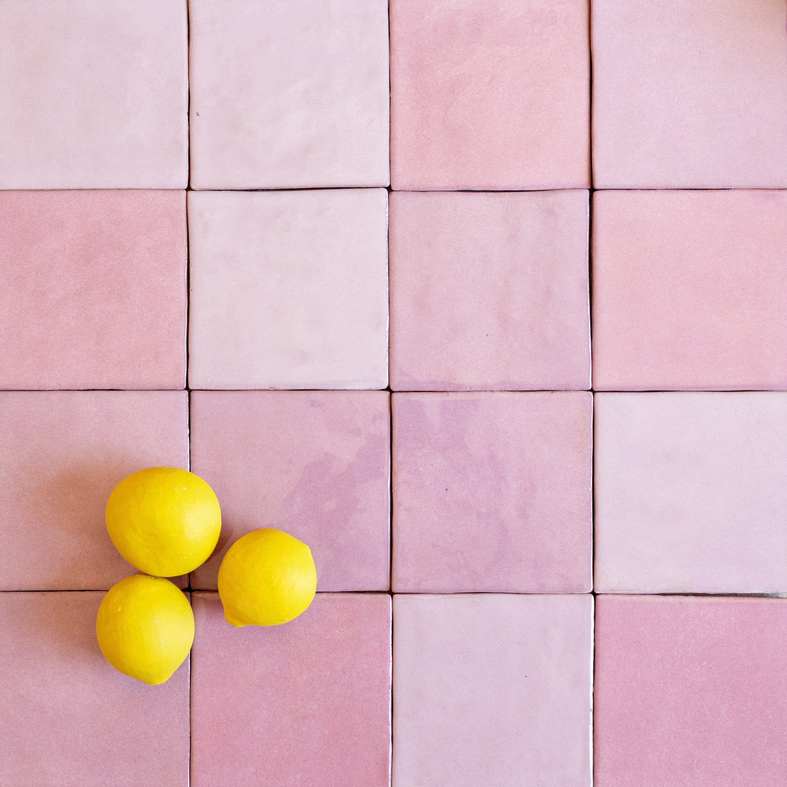 Square ceramic tiles in muted pink shades forming a grid with three bright yellow lemons arranged on the lower left corner