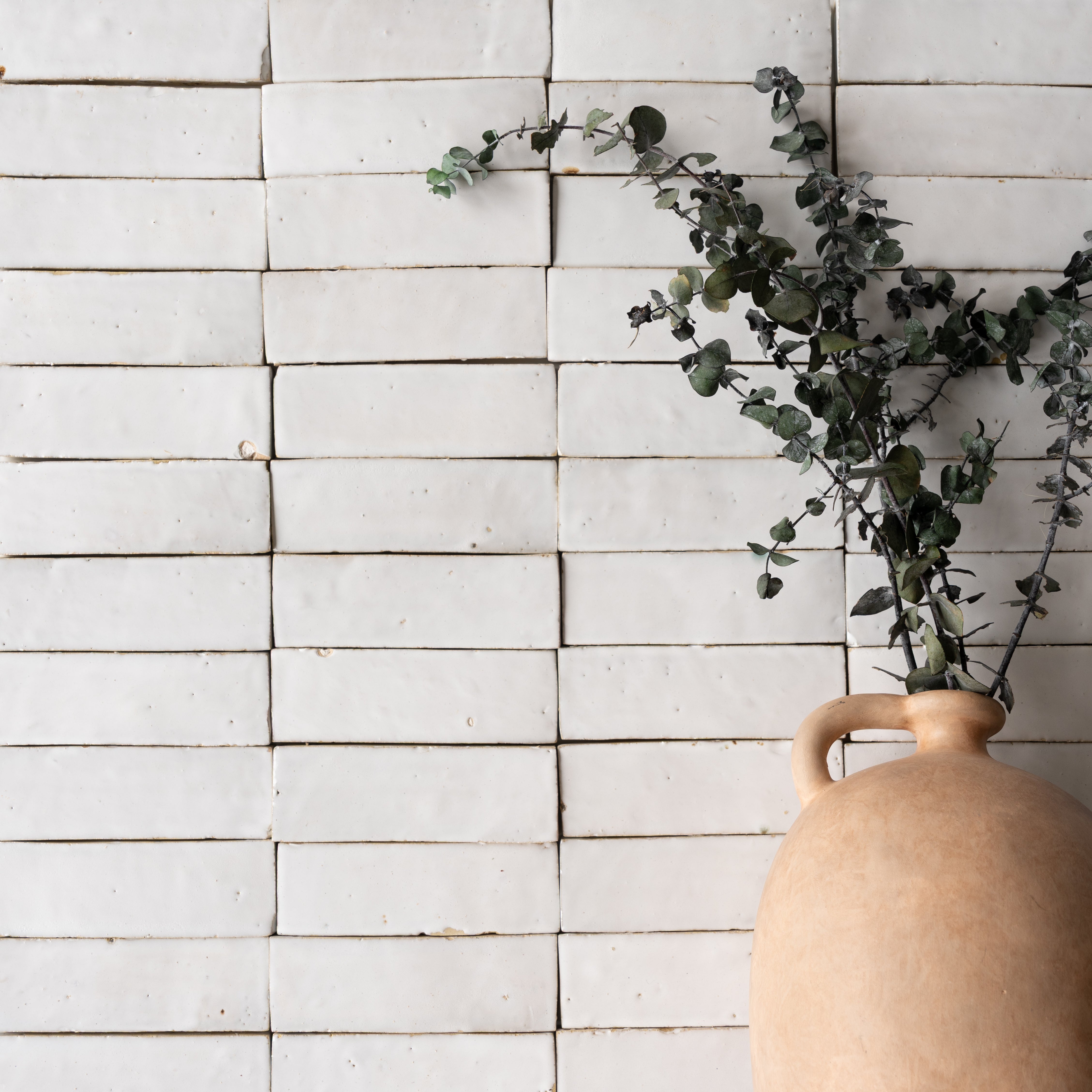Matte white ceramic zellige tiles arranged in a grid with a natural clay vase holding eucalyptus branches nearby