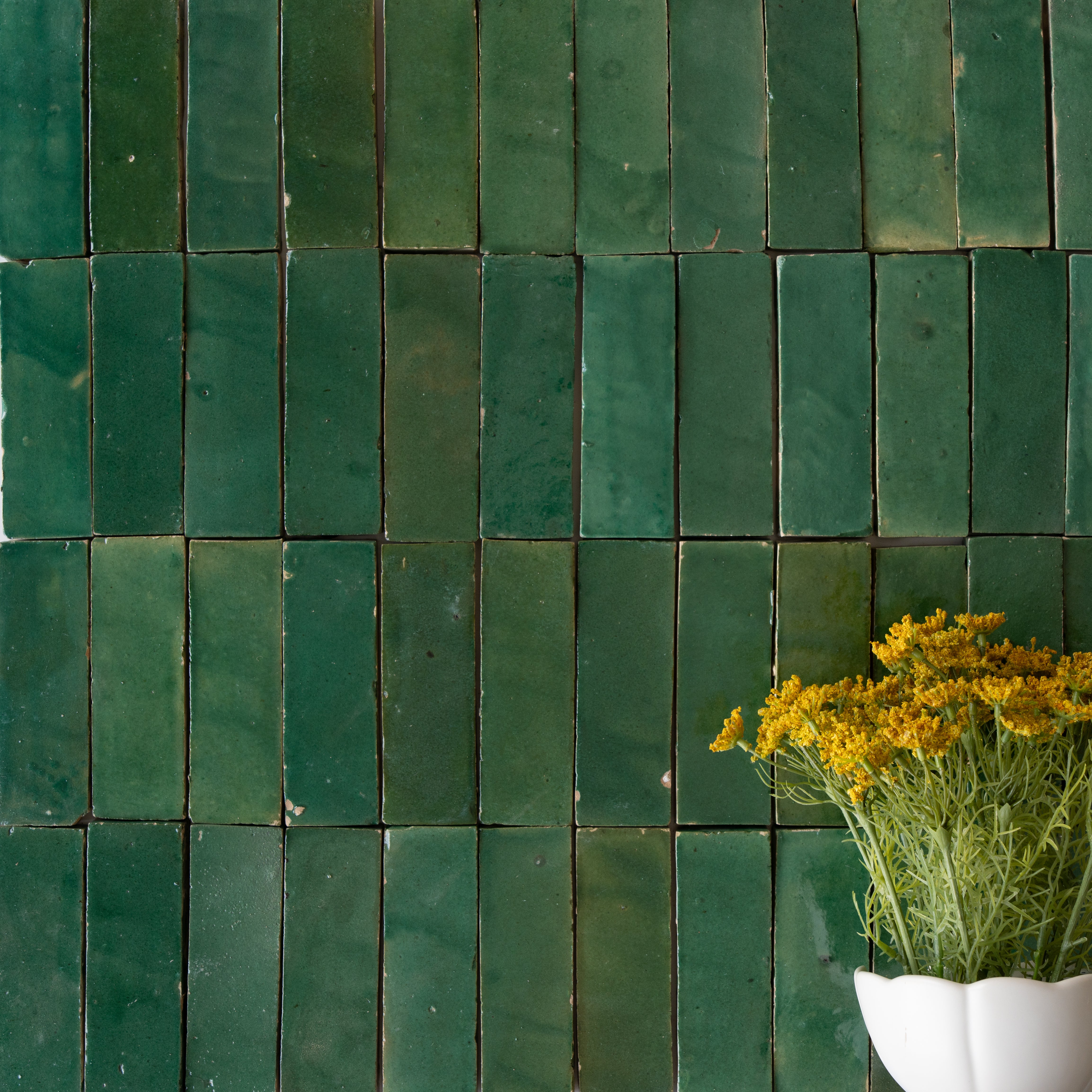 Glossy deep green ceramic zellige tiles with uneven surface texture beside a white vase holding yellow flowers