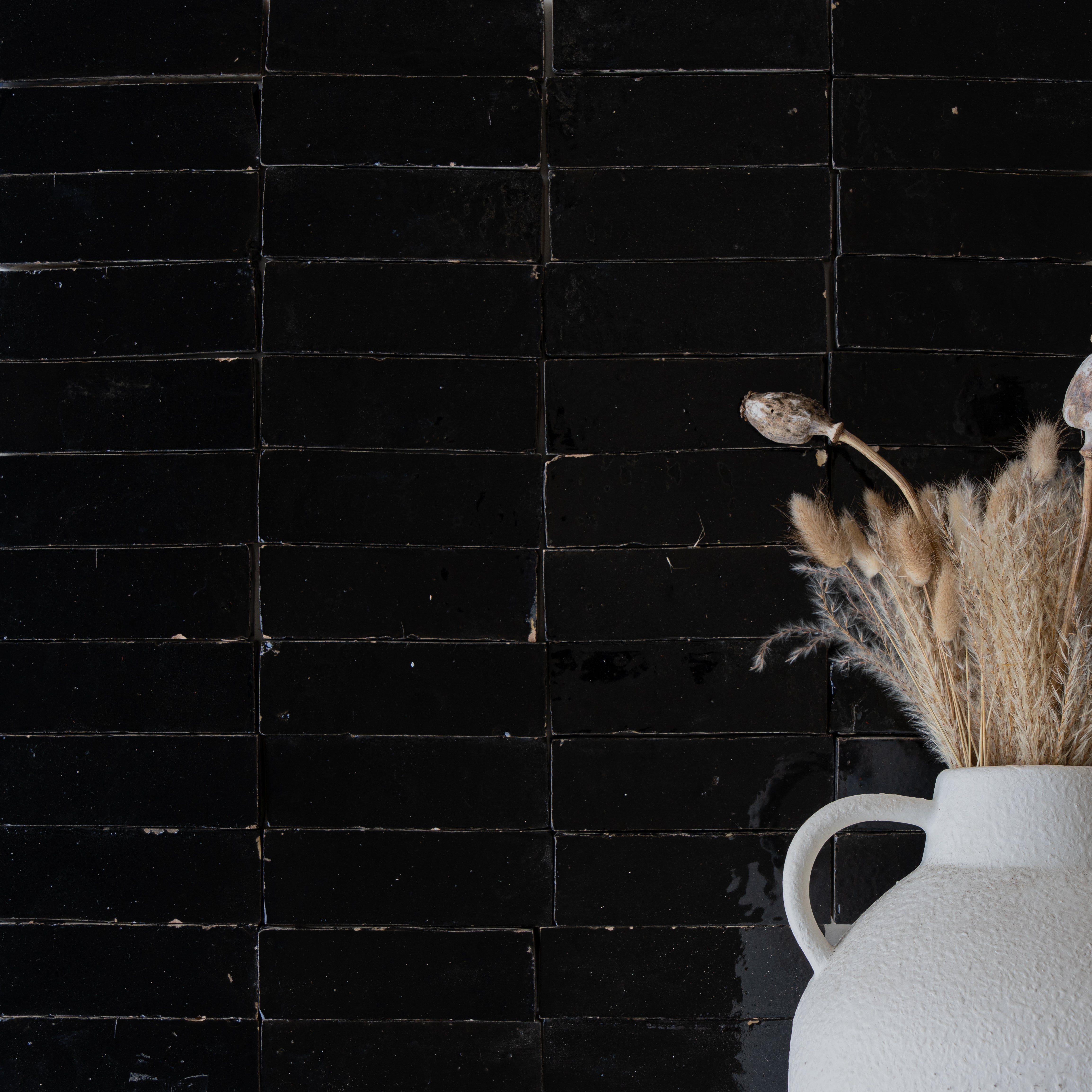 Glossy black ceramic zellige tiles with slight texture behind a white textured vase holding dried pampas and bunny tail grass