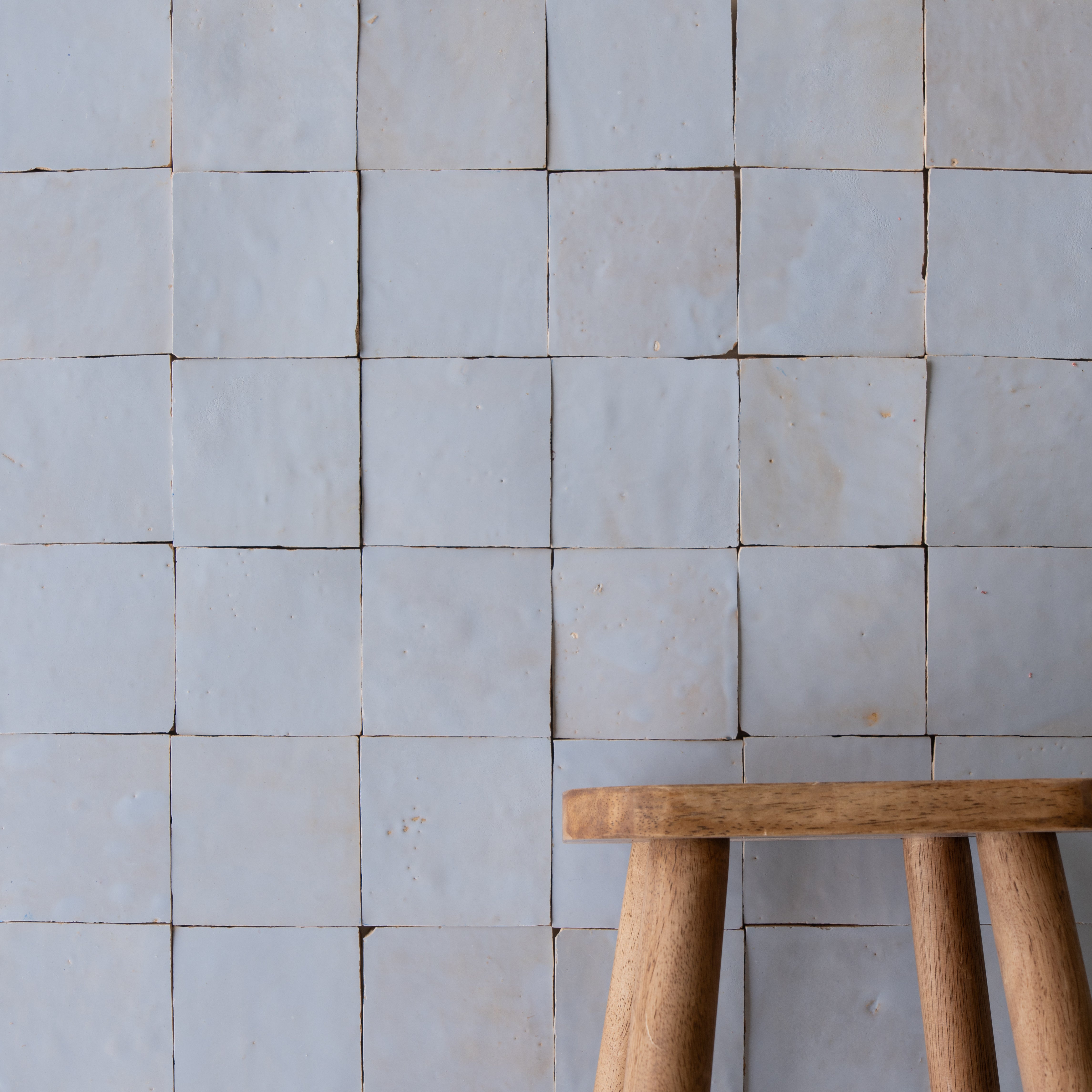 Soft matte light gray ceramic tiles forming a grid pattern with the edge of a natural wood stool visible in front