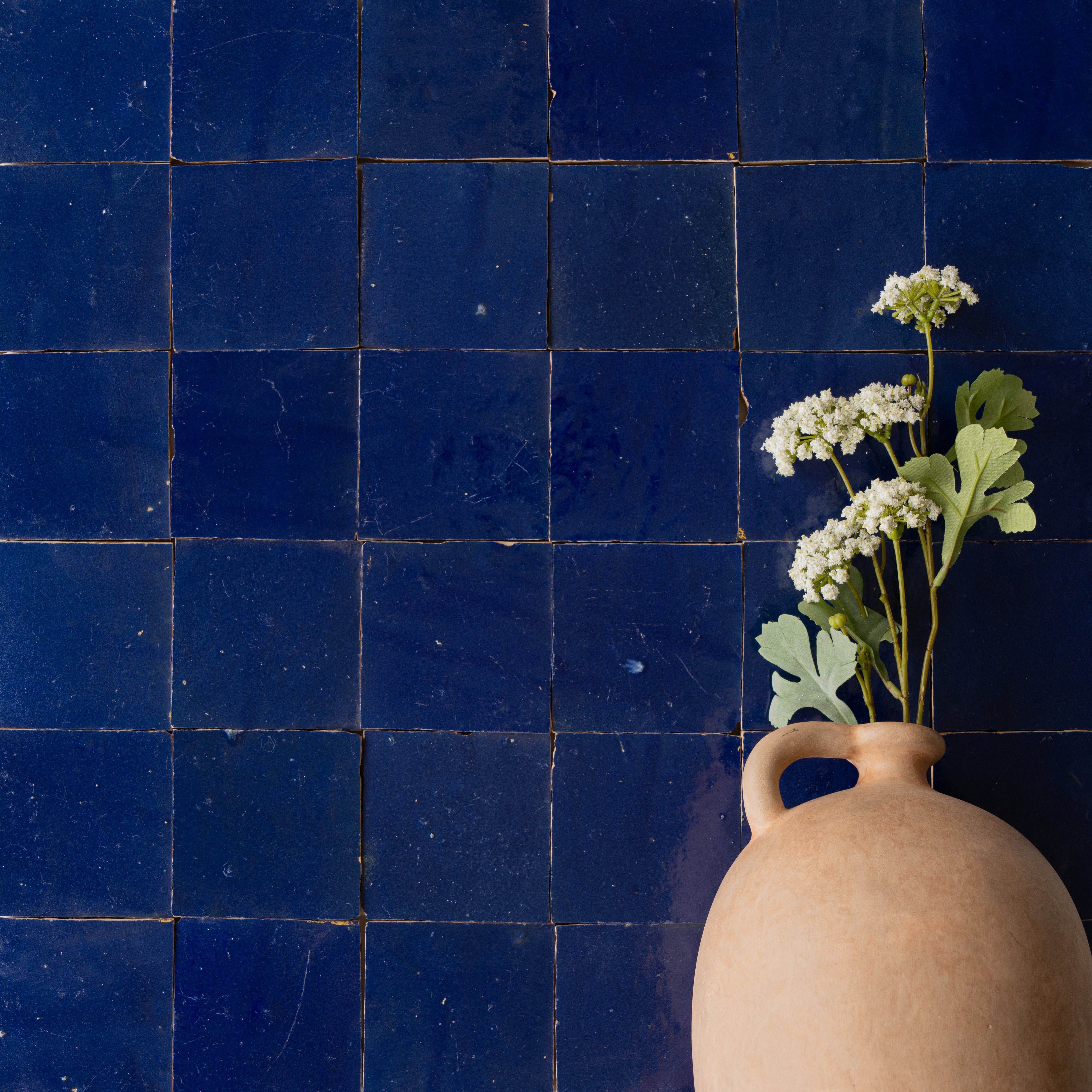 Glossy deep blue ceramic tiles with a slightly uneven surface paired with a simple beige clay vase holding white flowers