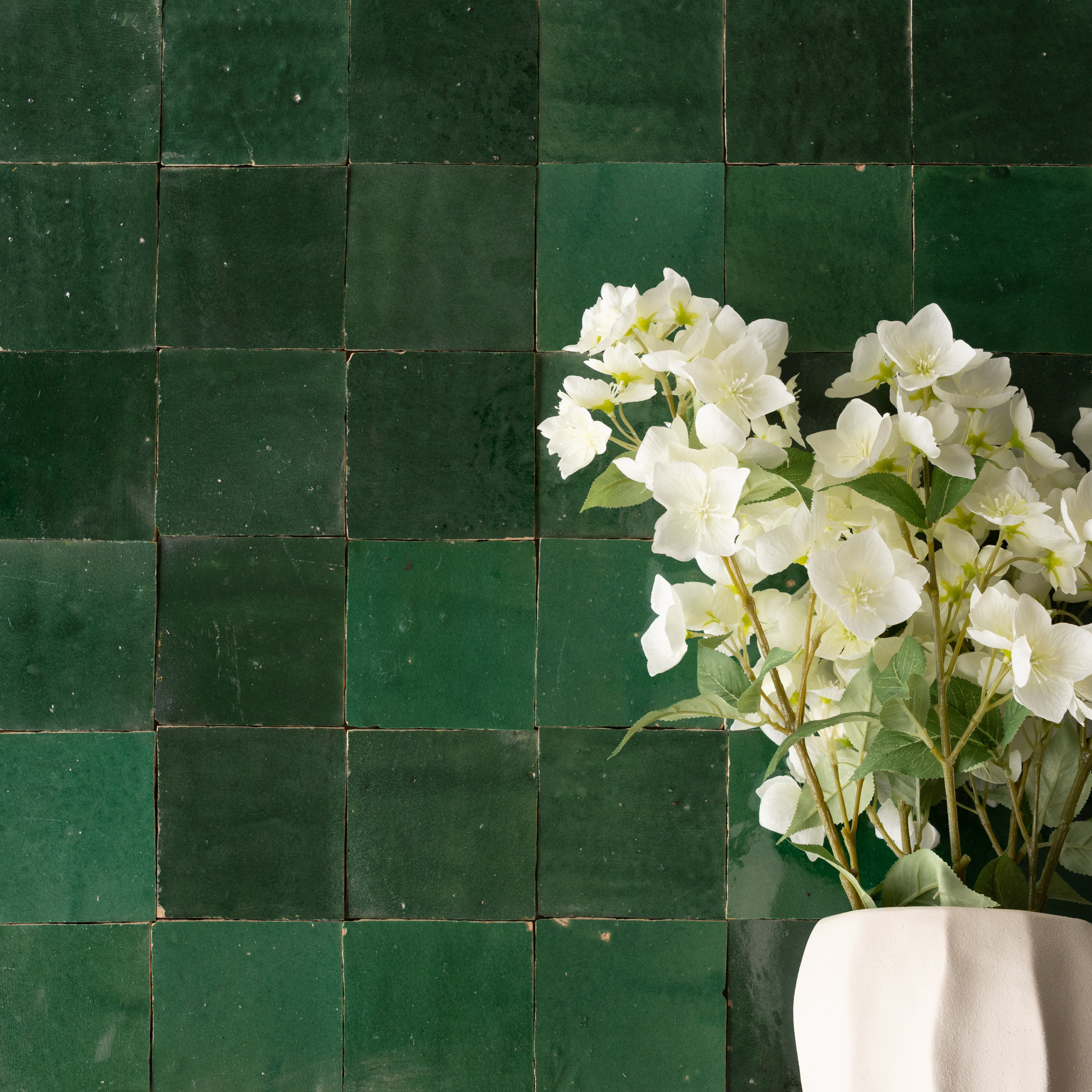 Glossy green ceramic zellige tiles with subtle texture and a white vase holding delicate white flowers on the right side