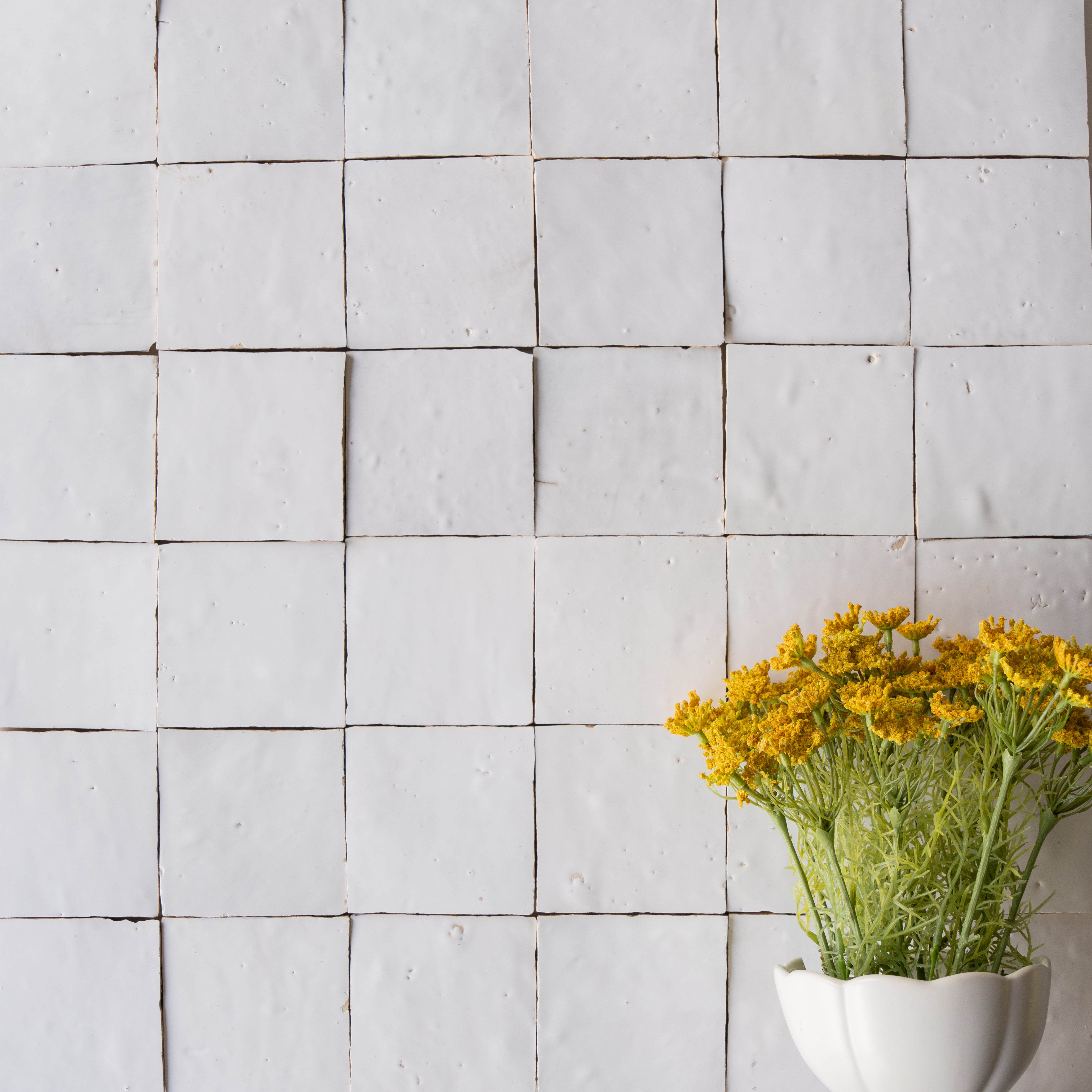 Matte off-white ceramic zellige tiles with subtle texture and a white vase holding bright yellow flowers at the corner