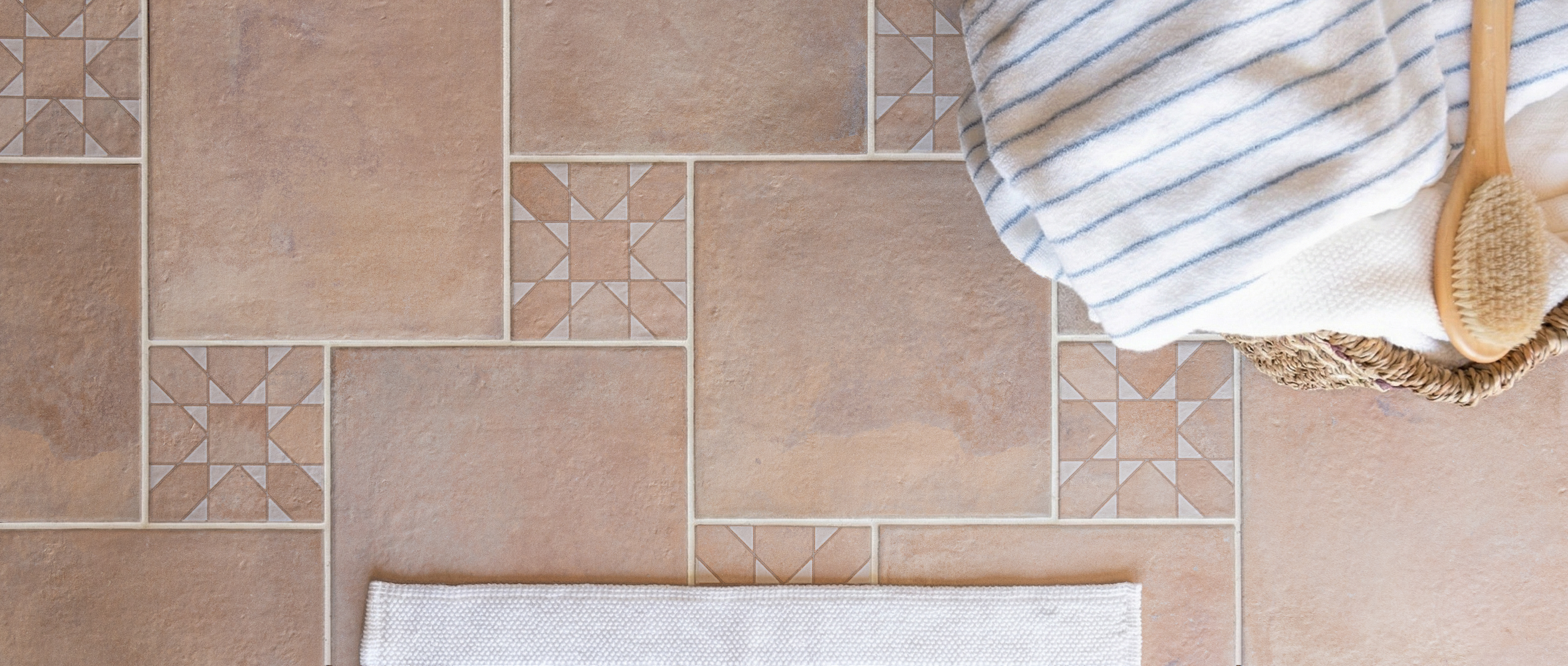 Overhead view of warm beige terracotta tiles with geometric insets next to a striped cloth and bath brush
